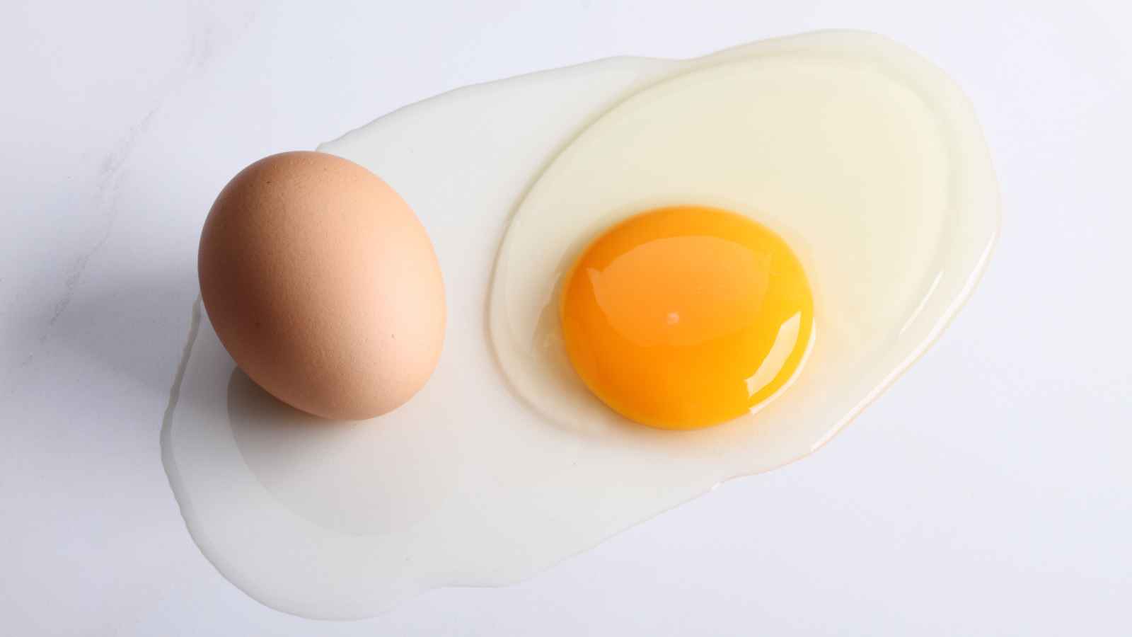 A whole brown egg sits next to a cracked egg with the yolk and egg white spread out on a white surface.
