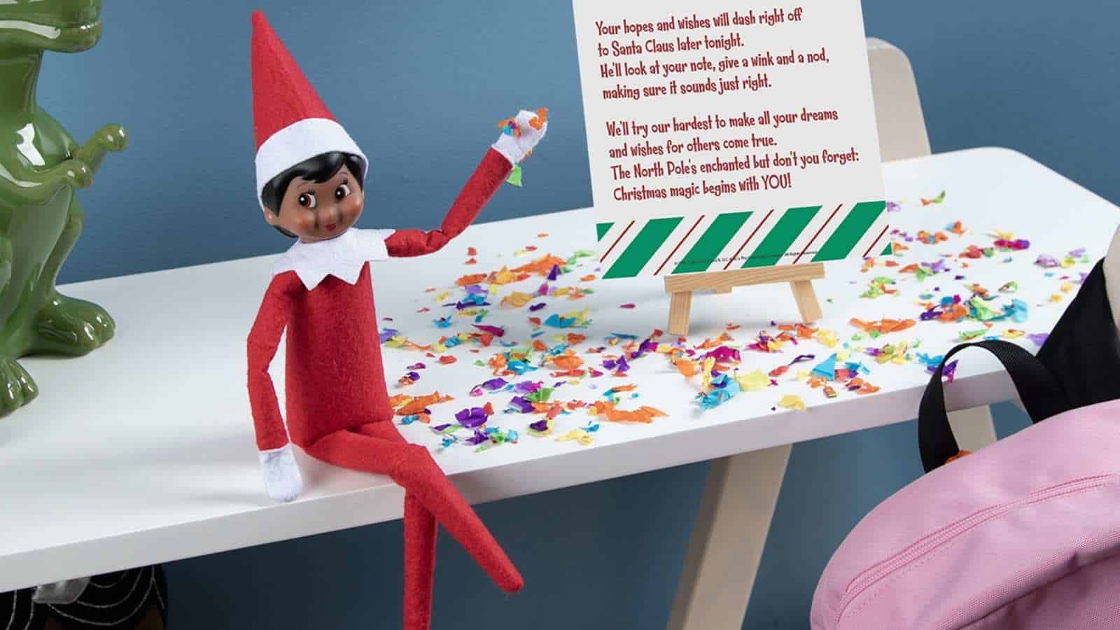 A red Elf on the Shelf doll sits on a white desk scattered with colorful confetti, next to a sign on an easel with text and a pink backpack.