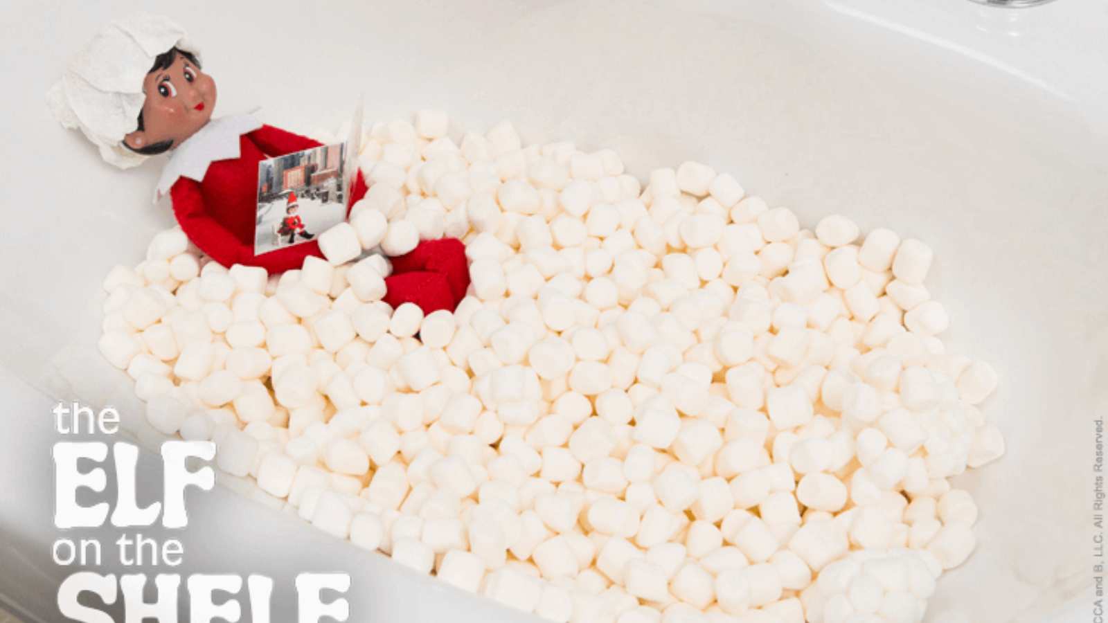An Elf on the Shelf doll sits in a bathtub filled with mini marshmallows, holding a small booklet.