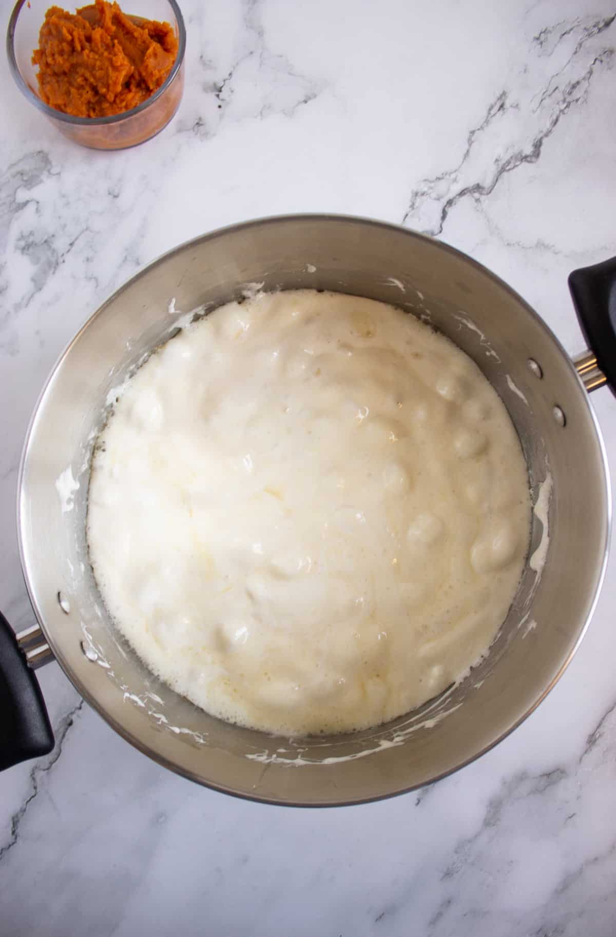 A saucepan with a bubbling white mixture on a marble countertop, with a small bowl of orange puree in the background.