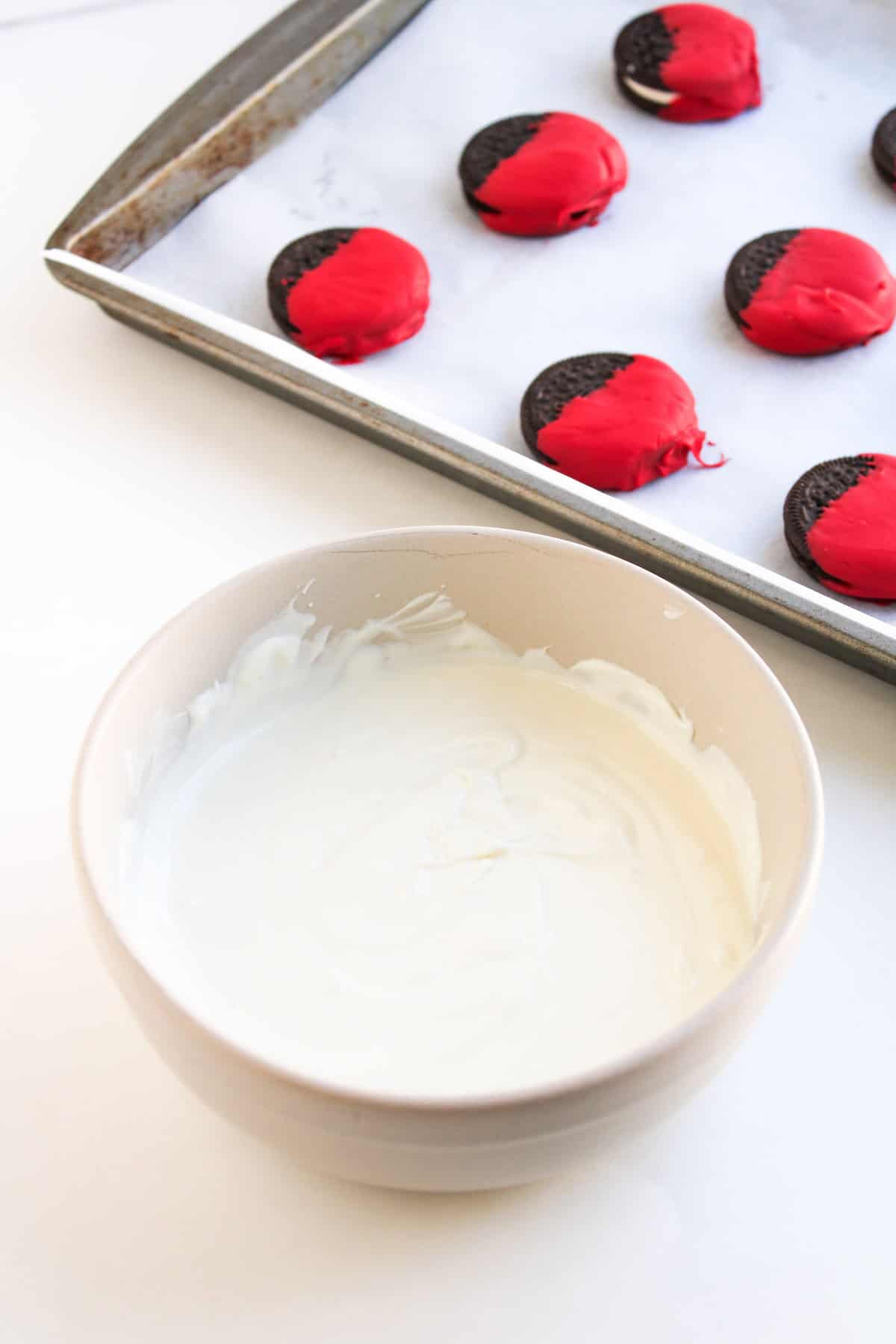 A bowl of melted white chocolate sits in the foreground, while a baking tray with Oreos half-dipped in red coating is in the background.