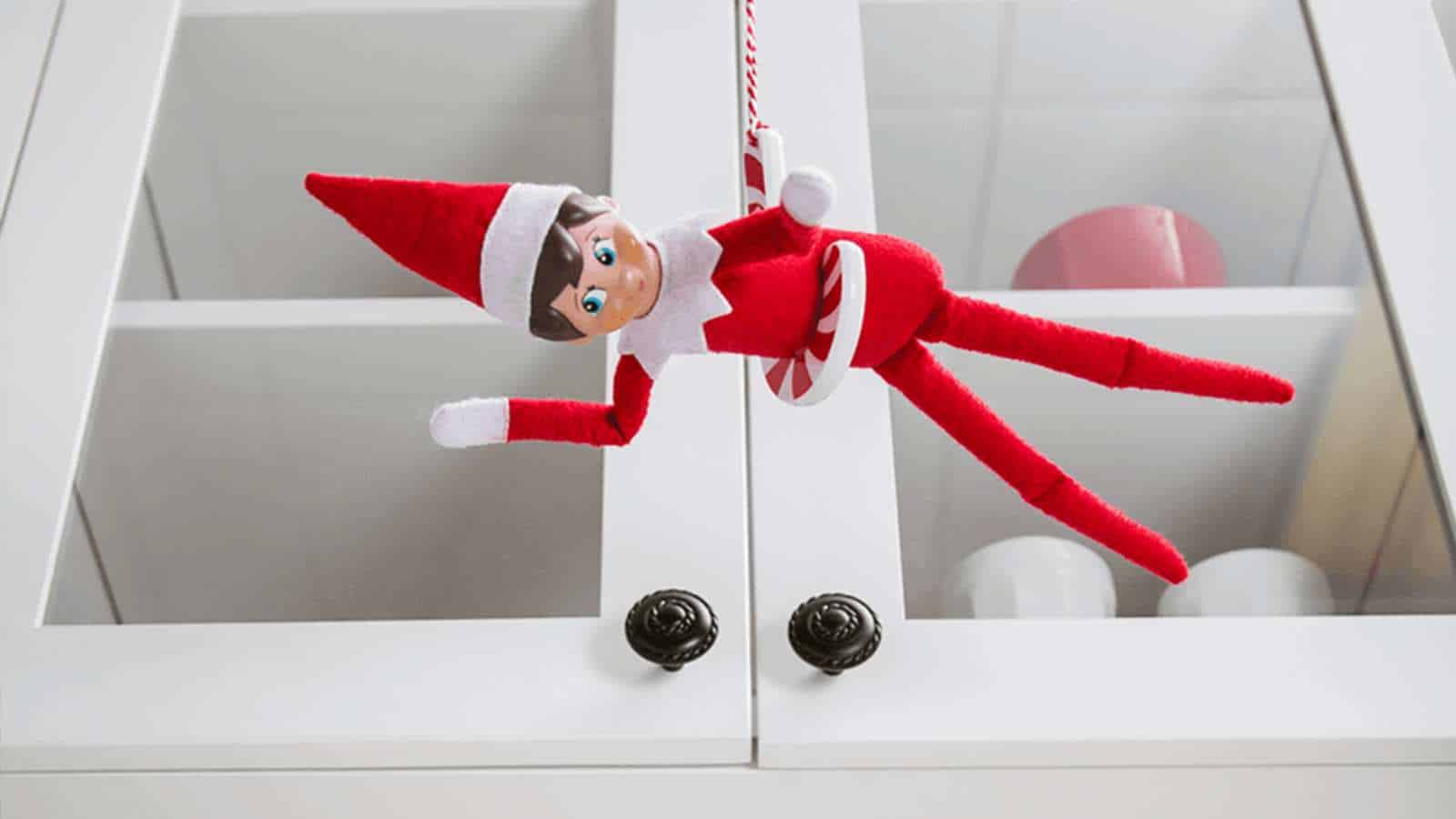 A red and white Elf on the Shelf doll hangs from a candy cane ornament in front of white cabinet doors with glass panels.
