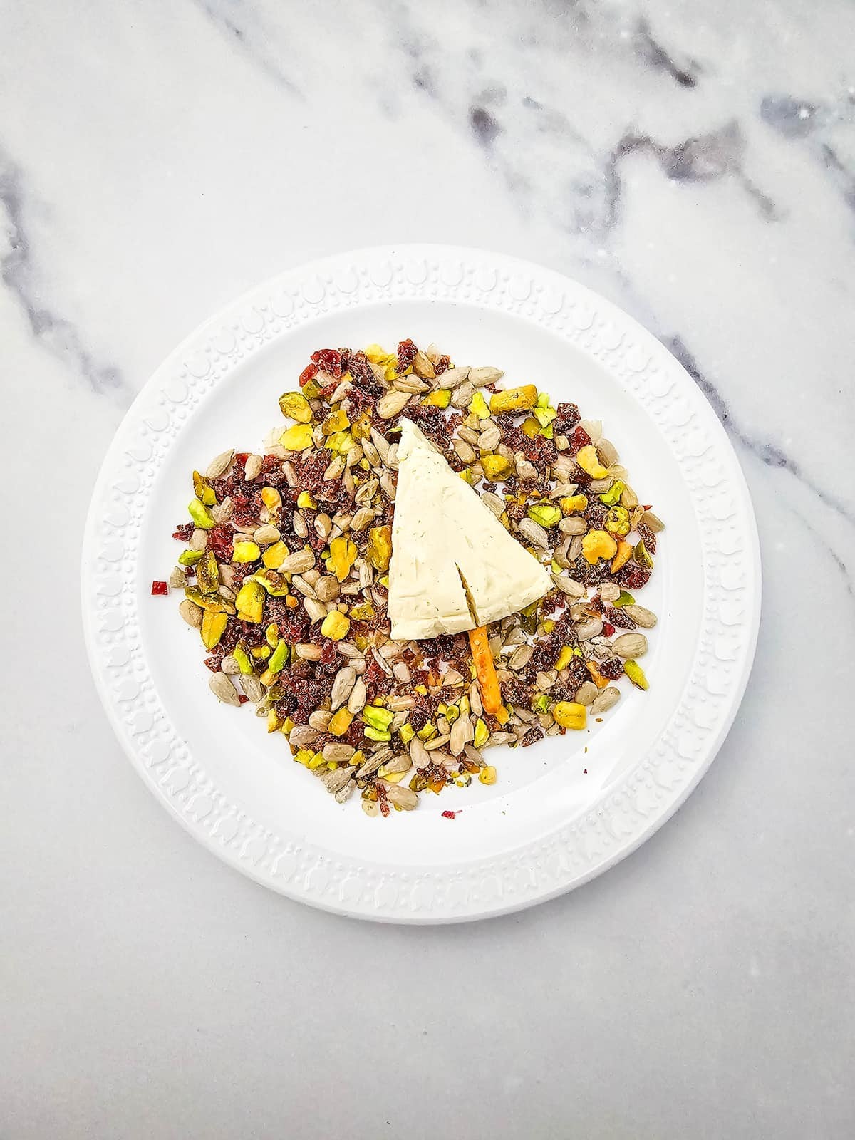 A wedge of cheese sits on a white plate, surrounded by a mix of dried fruits and seeds, on a marble surface.