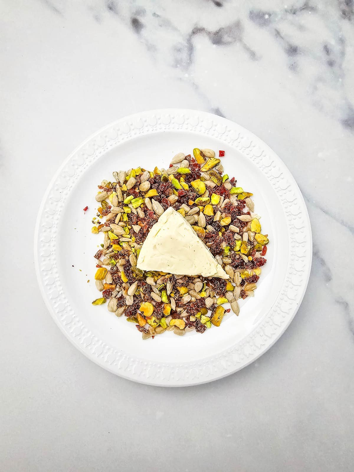 A slice of cheese sits on a mix of dried fruit, seeds, and nuts, arranged on a white plate atop a light gray marble surface.