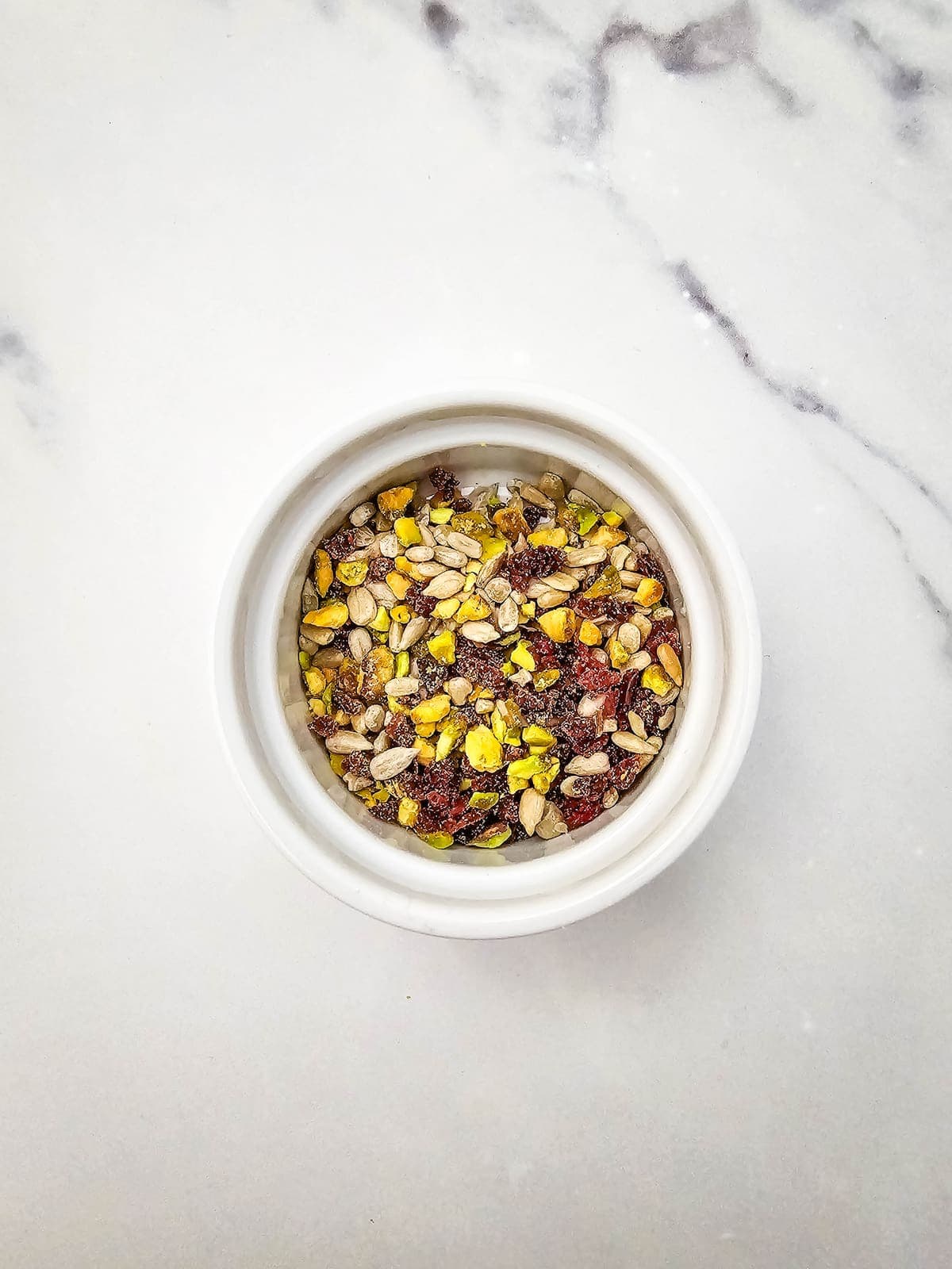 A white bowl filled with a mix of seeds and grains sits on a light-colored marble surface.