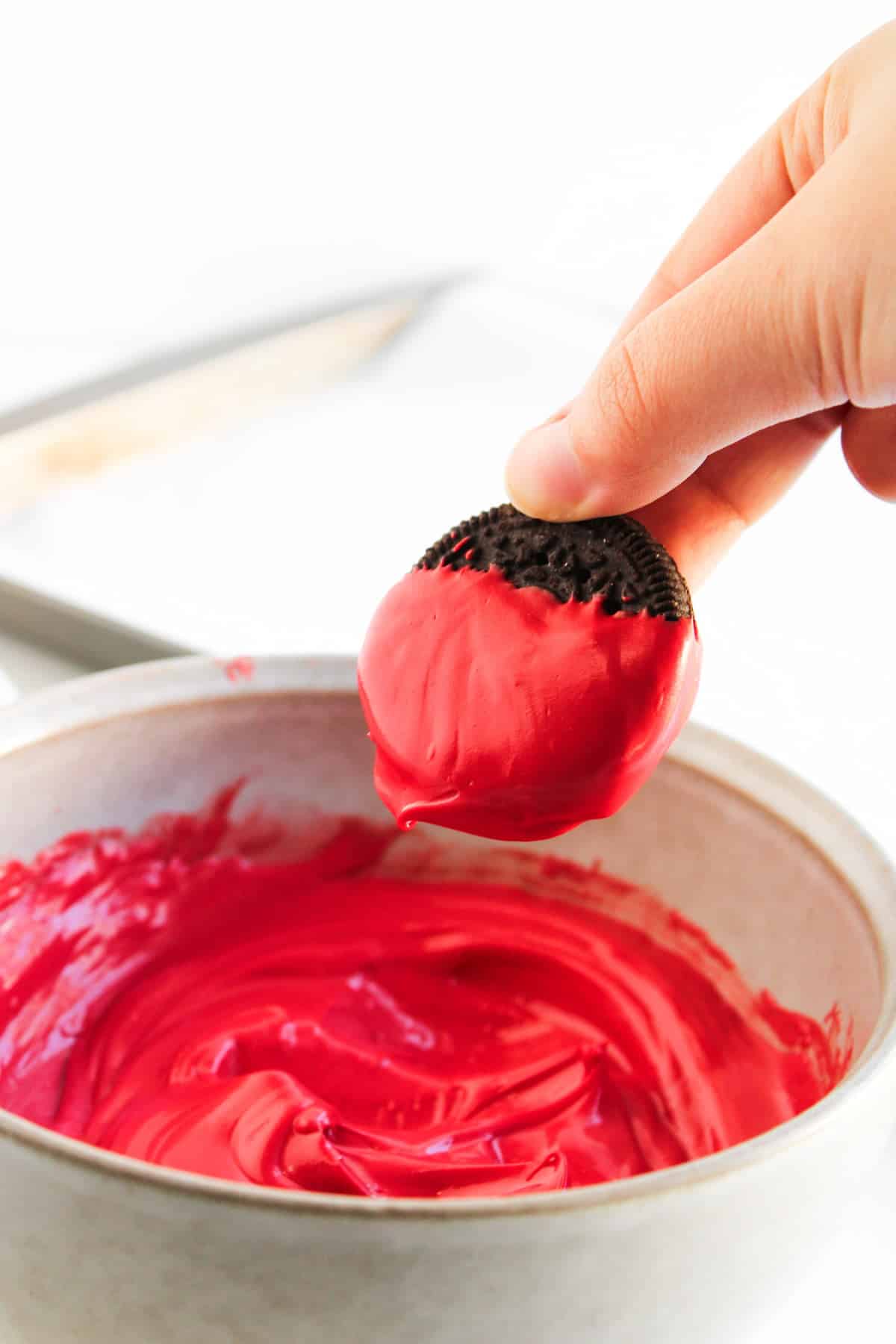 A hand dips an Oreo cookie halfway into a bowl of melted red chocolate.