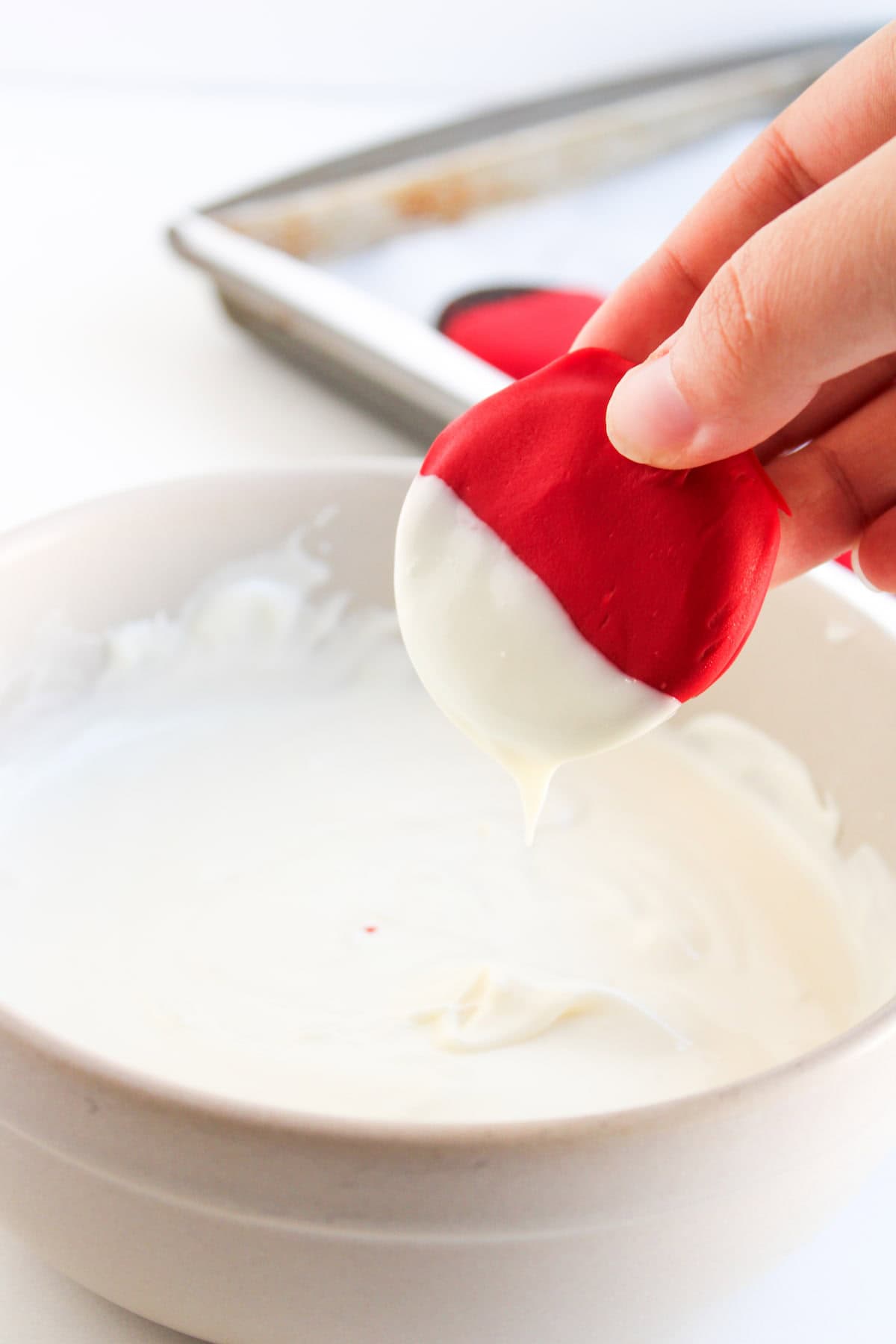 A hand dips a red item, possibly a cookie, into a bowl of melted white chocolate, with a baking tray in the background.