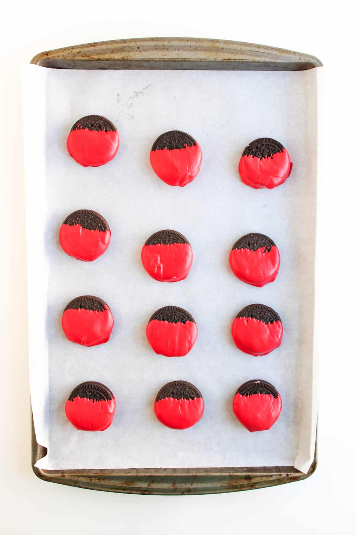 A baking tray lined with parchment paper holds twelve chocolate cookies dipped halfway in red coating, arranged in rows.