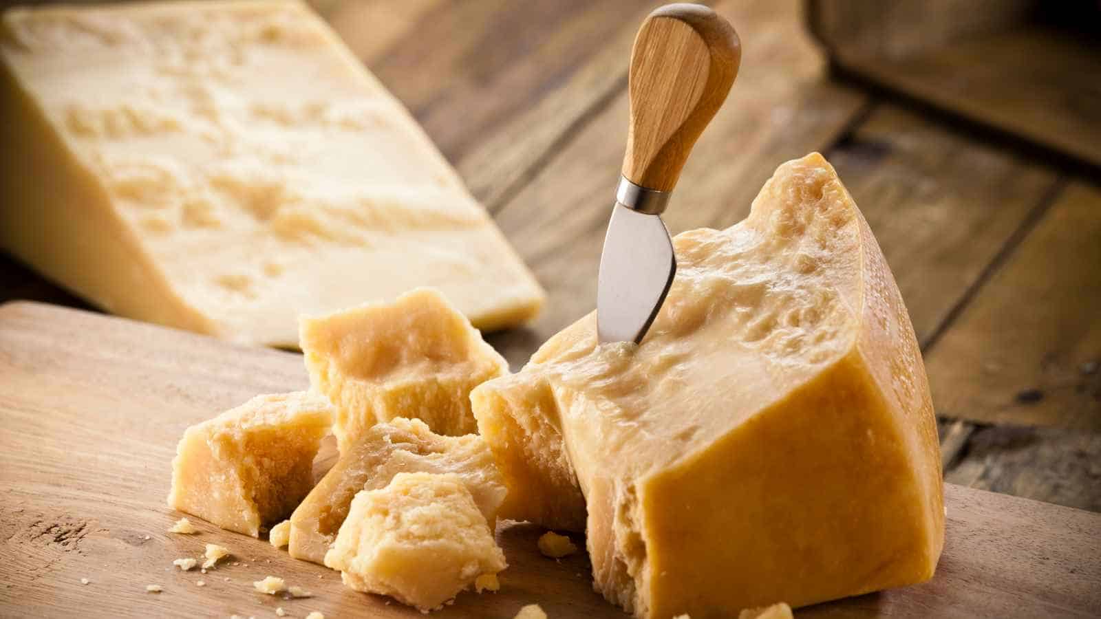 A wedge of hard cheese with a cheese knife inserted, surrounded by several broken chunks, on a wooden cutting board.