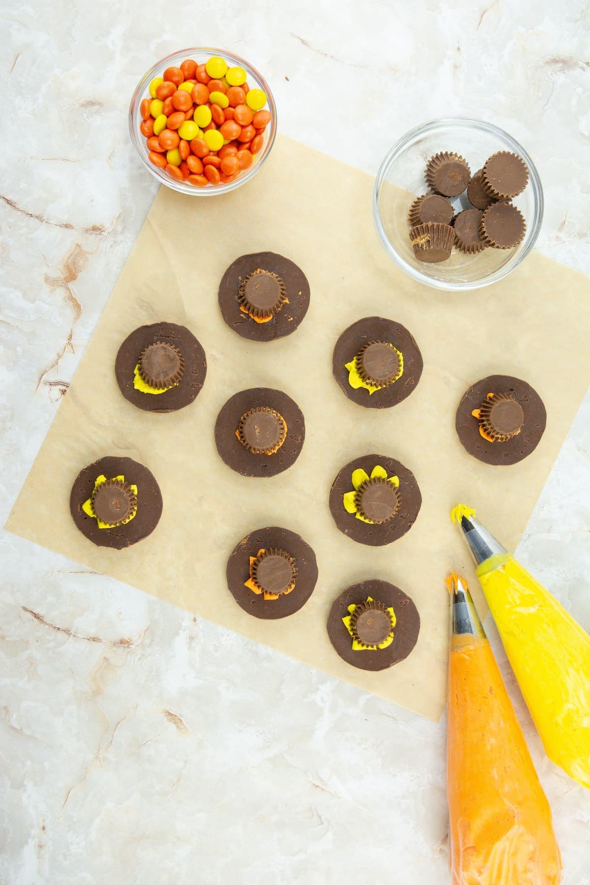 Chocolate cookies topped with mini peanut butter cups are arranged on parchment paper, surrounded by bowls of candies and piping bags filled with orange and yellow frosting.