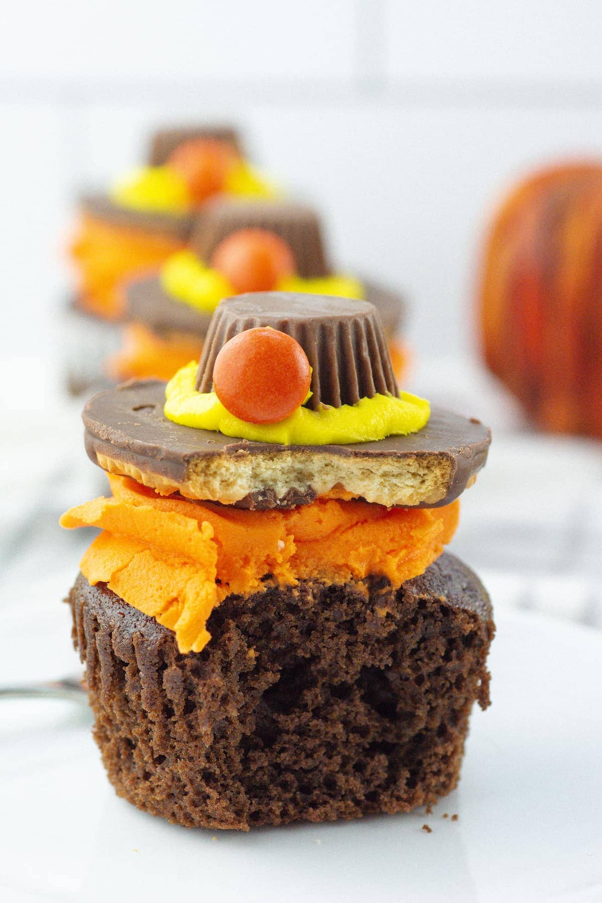 A chocolate cupcake with orange frosting is topped with a cookie, a chocolate peanut butter cup, yellow icing, and a round orange candy. A bite has been taken from the cupcake.