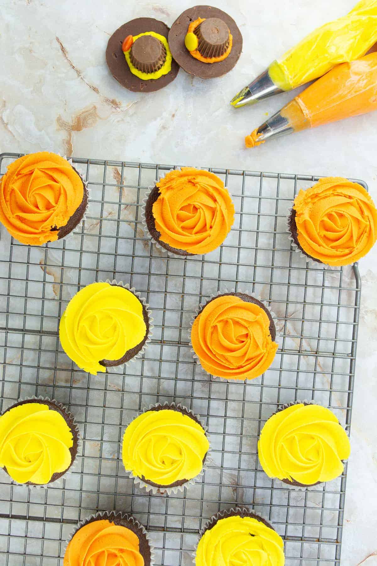 Cupcakes with orange and yellow frosting on a cooling rack, with two frosted cupcakes and icing bags nearby on a light countertop.