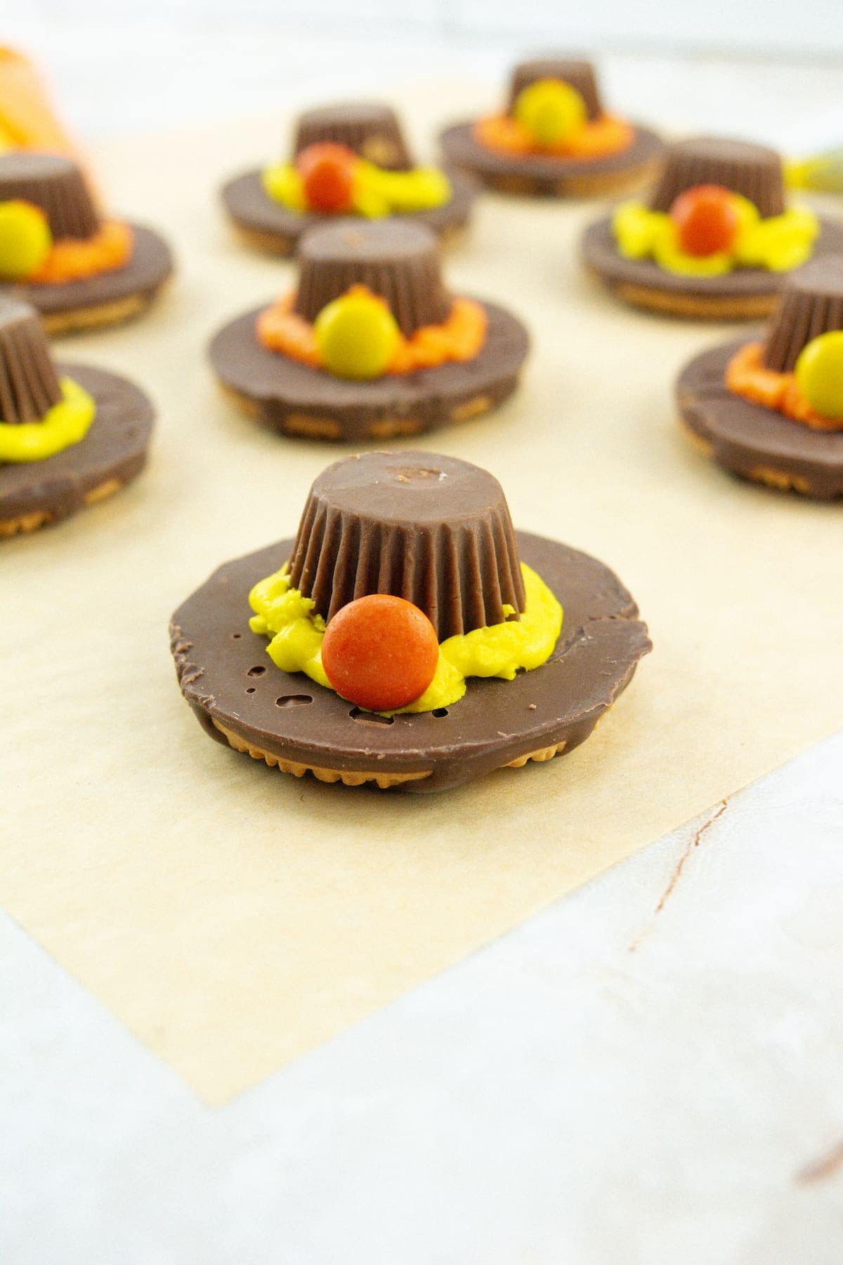Several chocolate treats resembling pilgrim hats sit on parchment paper, made from cookies, peanut butter cups, frosting, and colorful candy pieces.