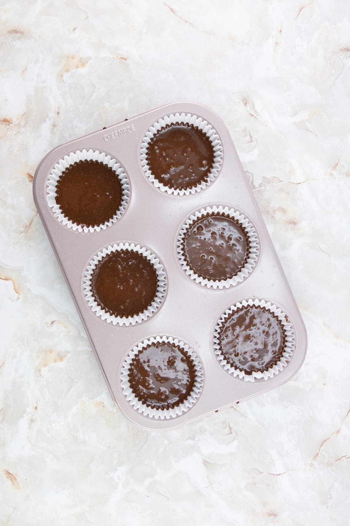 A muffin tin with six paper liners, each filled with chocolate batter, sits on a light marble countertop.