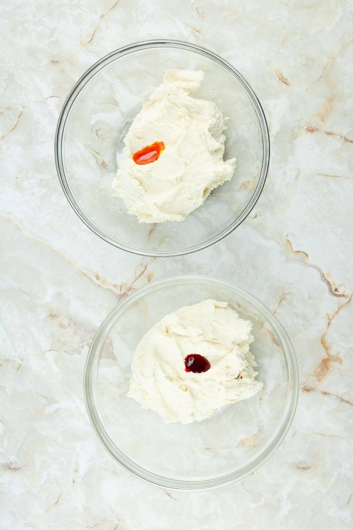Two glass bowls each containing a scoop of white cream cheese, one with an orange drop and one with a yellow drop of food coloring, on a light marble surface.