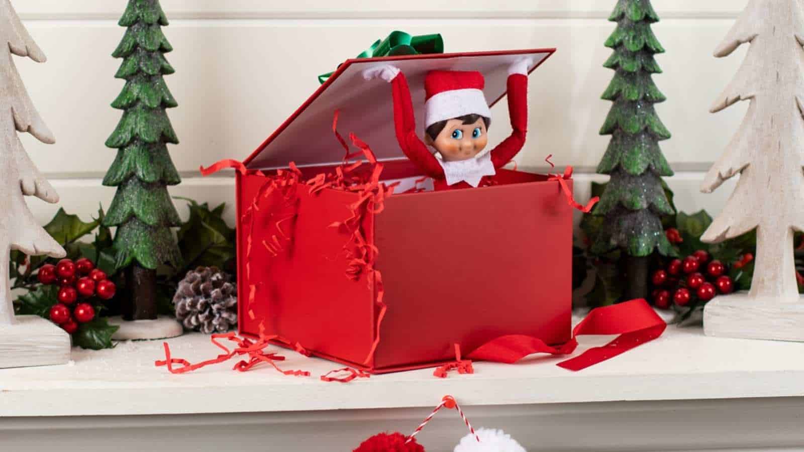 A toy elf wearing a red and white outfit pops out of an open red gift box, surrounded by festive decorations and artificial trees.