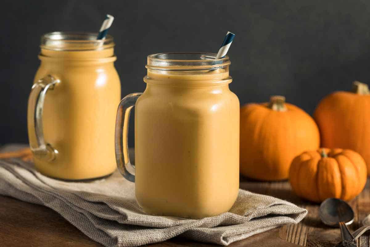 Two mason jars filled with orange smoothies, each with a striped straw, sit on a cloth napkin next to small pumpkins on a wooden surface—perfect for enjoying after learning how to freeze pumpkin for fresh smoothies anytime.