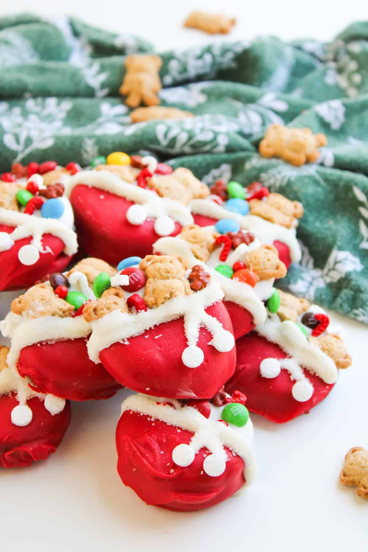 A pile of red, Santa's toy bag dipped cookies decorated with bear-shaped cookies, white icing, and colorful candies sits on a white surface with a green patterned cloth in the background.