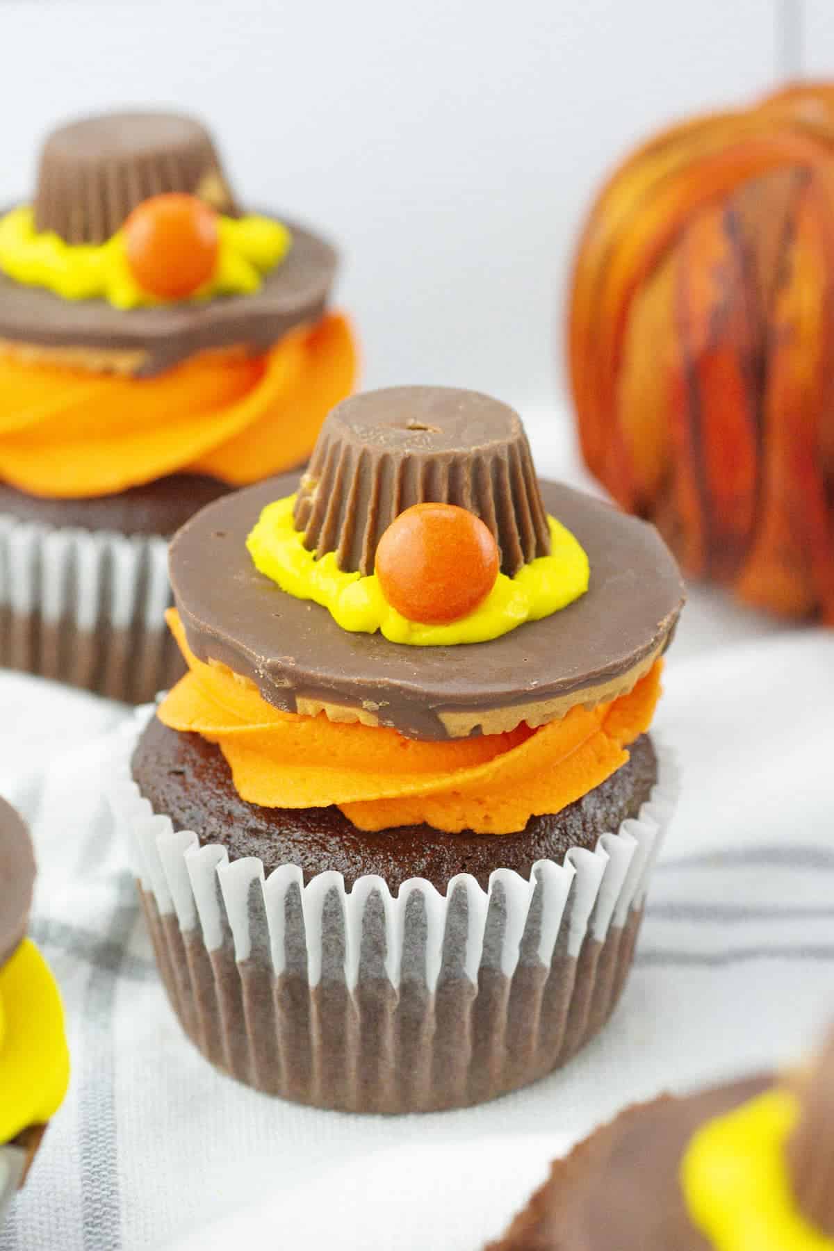 Chocolate cupcake with orange and yellow frosting, topped with a chocolate hat made from a peanut butter cup and orange candy. A decorative pumpkin is in the background.