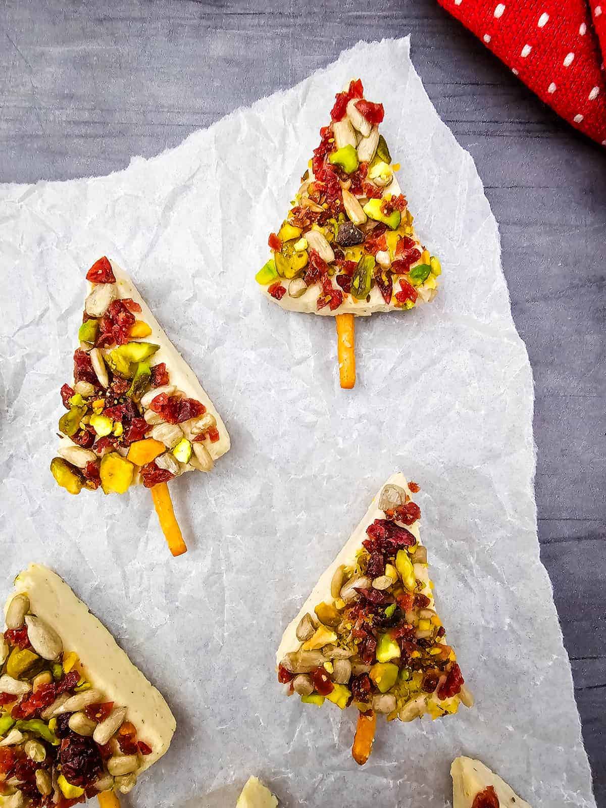 Triangle-shaped treats decorated with nuts and dried fruit to resemble Christmas trees, each with a pretzel stick as the tree trunk, arranged on parchment paper.