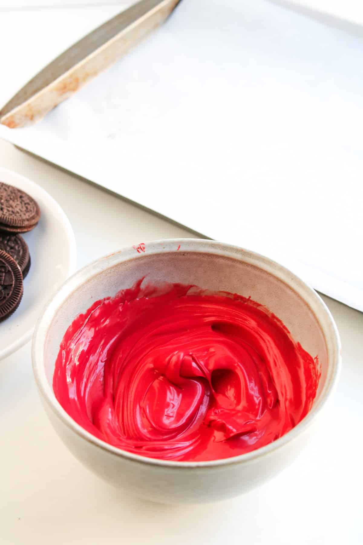 A bowl of smooth, bright red melted chocolate sits on a white surface next to a plate of Oreos and a baking sheet lined with parchment paper.