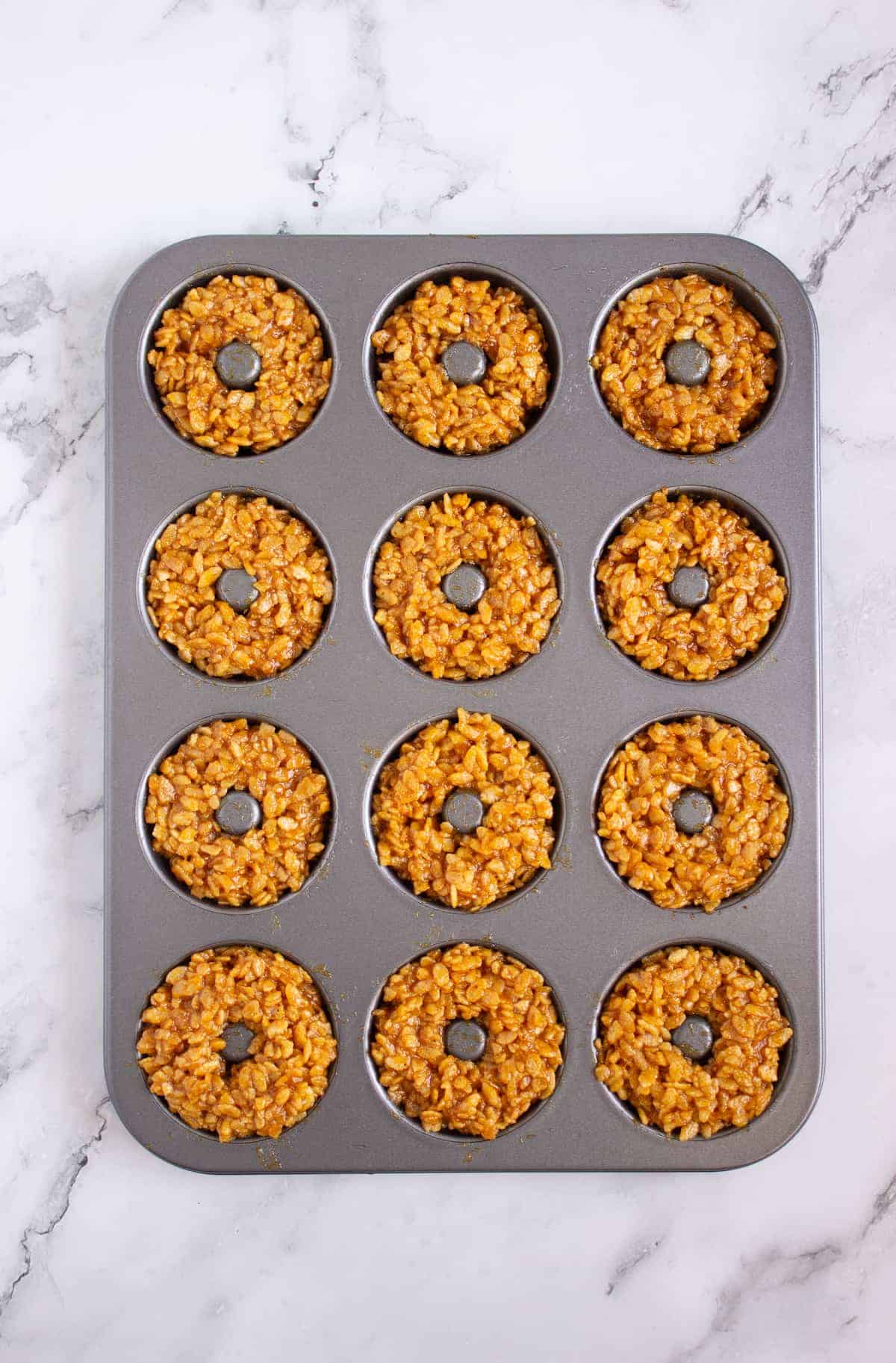 A muffin tin with twelve circular compartments, each filled with a sticky, caramel-colored rice mixture shaped like donuts, sits on a marble countertop.