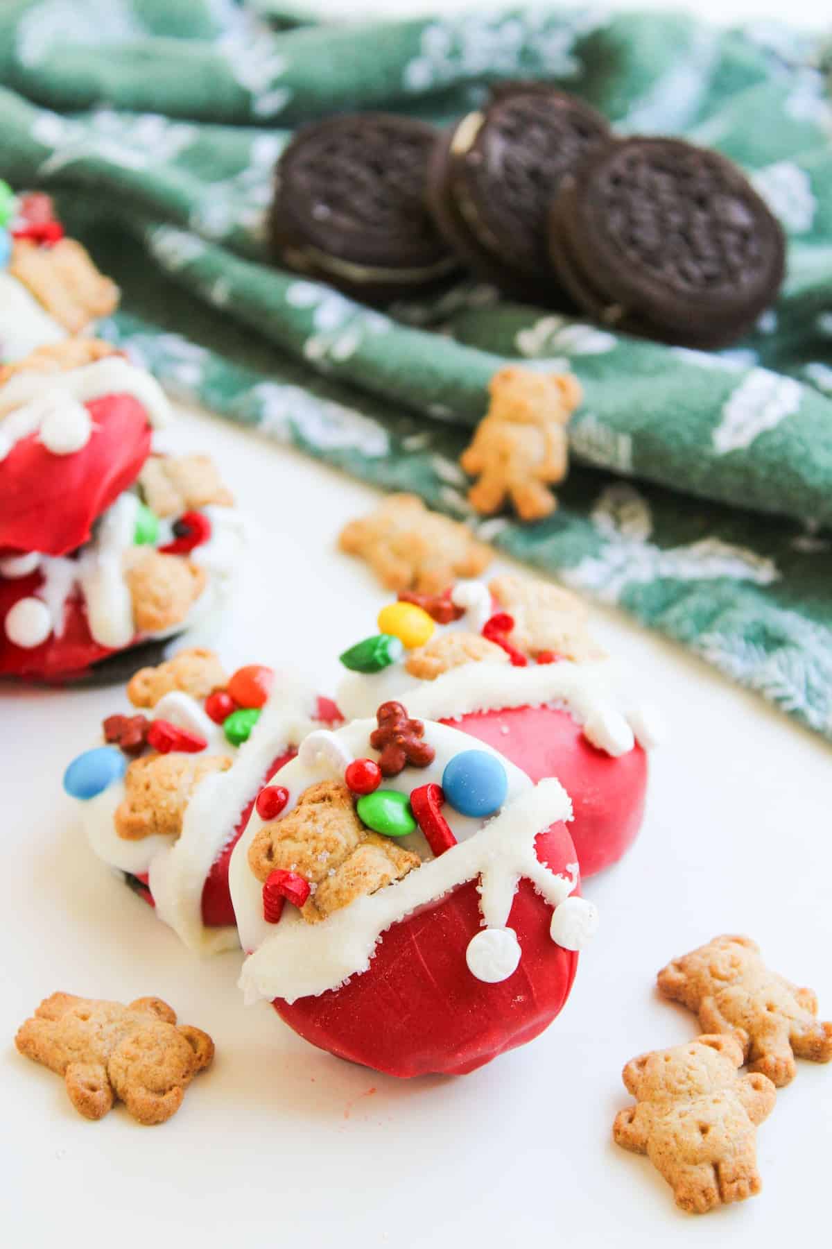 Three red, round holiday treats decorated with white icing, candy pieces, mini Santa's toy bag cookies, and colorful sprinkles, with Oreos and a green cloth in the background.