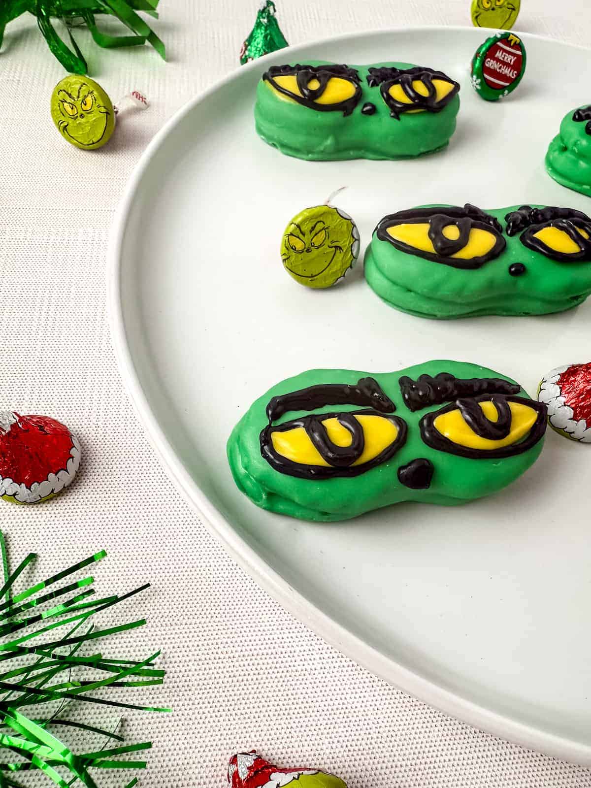 A nutter butter cookies decorated with melted green chocolate and yellow eyes resembling the Grinch are arranged on a white plate, surrounded by Grinch-themed candy and holiday decorations.