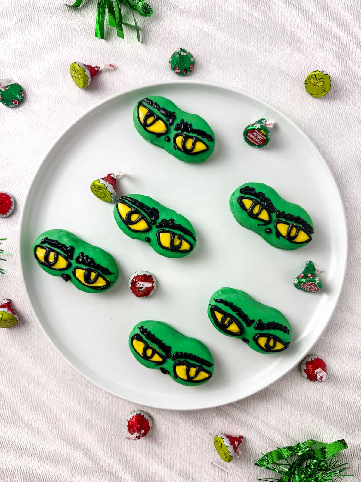 A white plate with six nutter butter cookies decorated with yellow and black eyes, resembling the Grinch, surrounded by holiday candy and decorations.