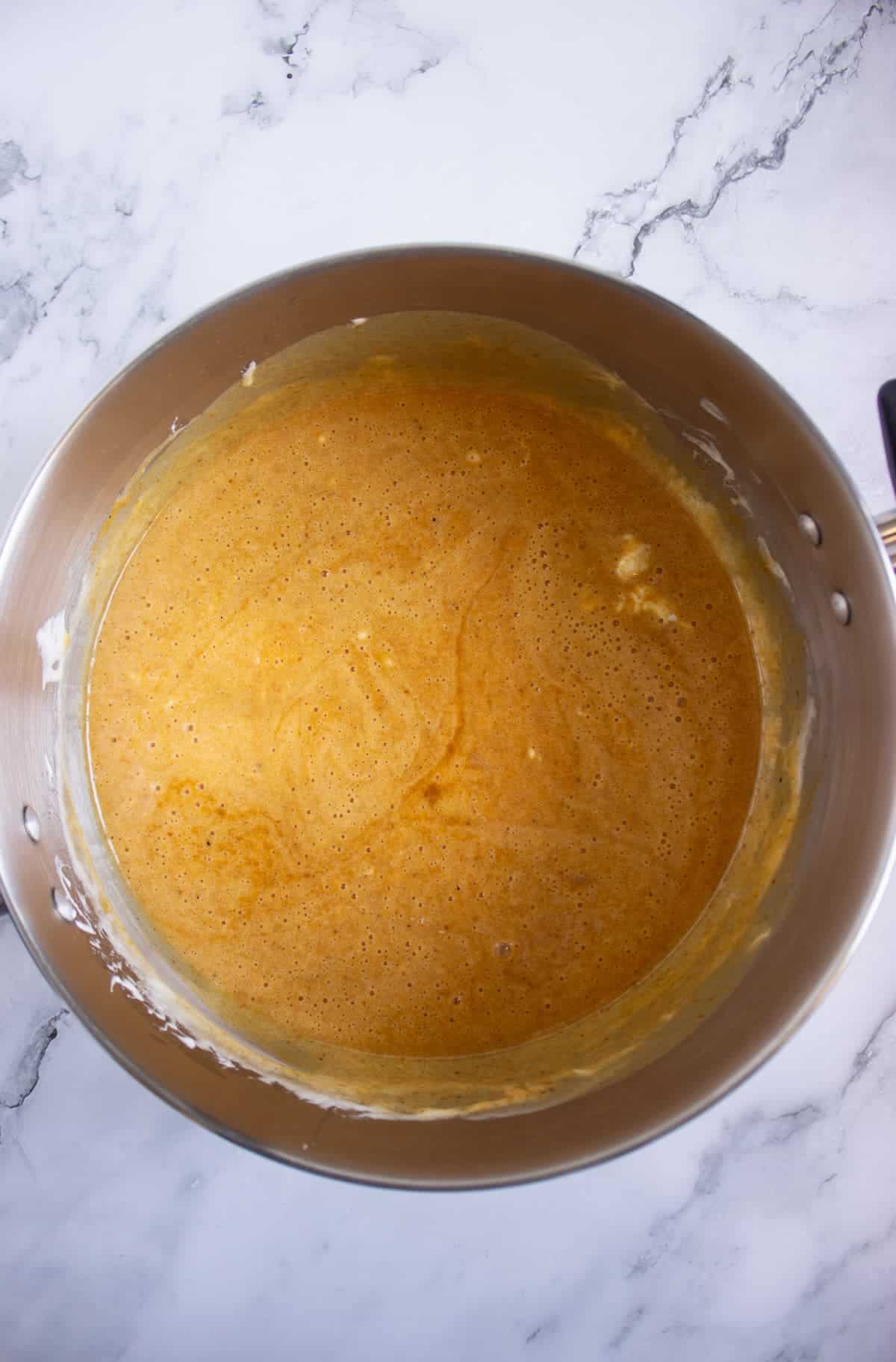 A metal mixing bowl filled with a light brown batter mixture, placed on a white marble surface.