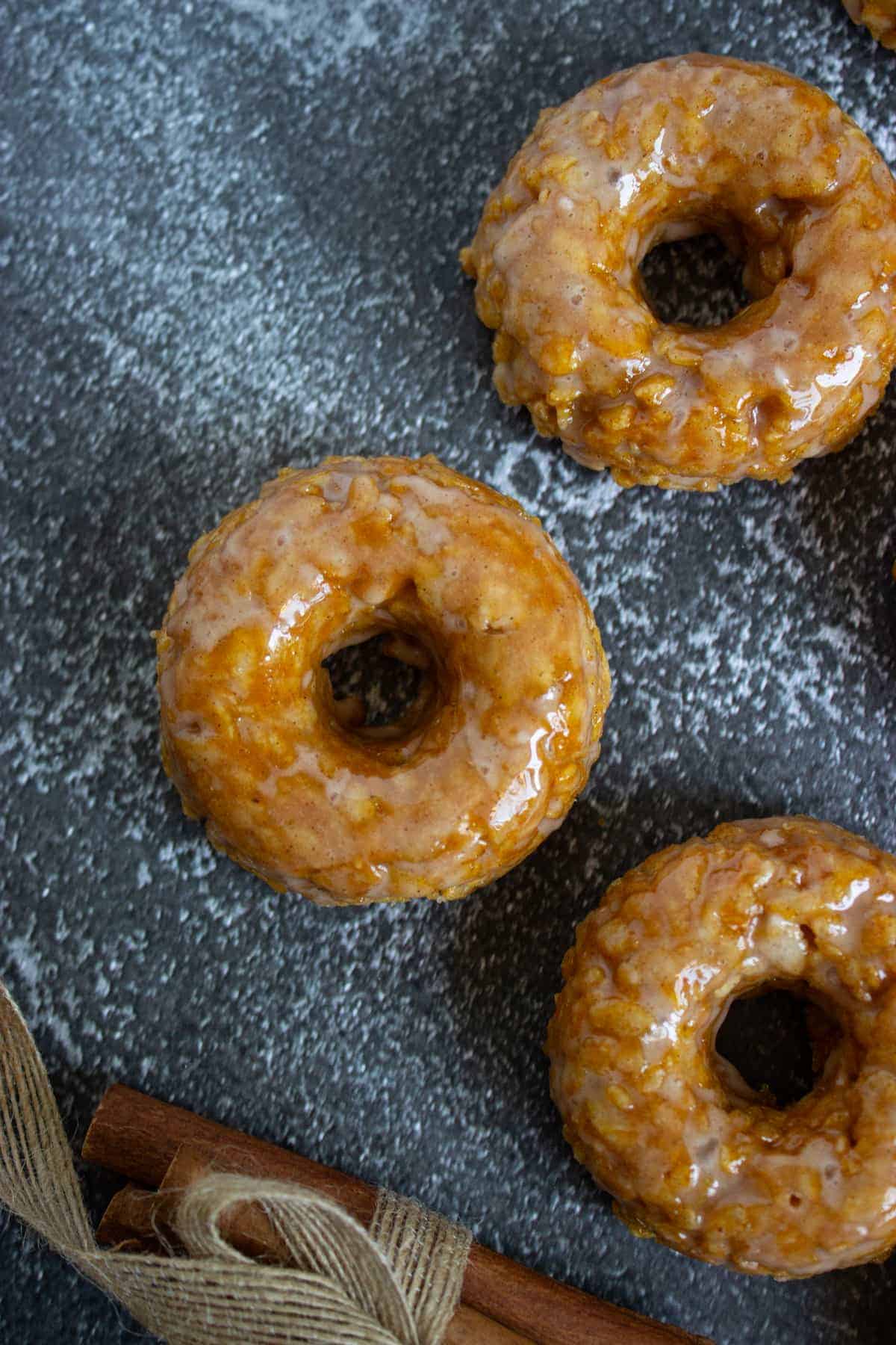 Three glazed donuts are arranged on a dark, textured surface near a bundle of cinnamon sticks tied with twine.
