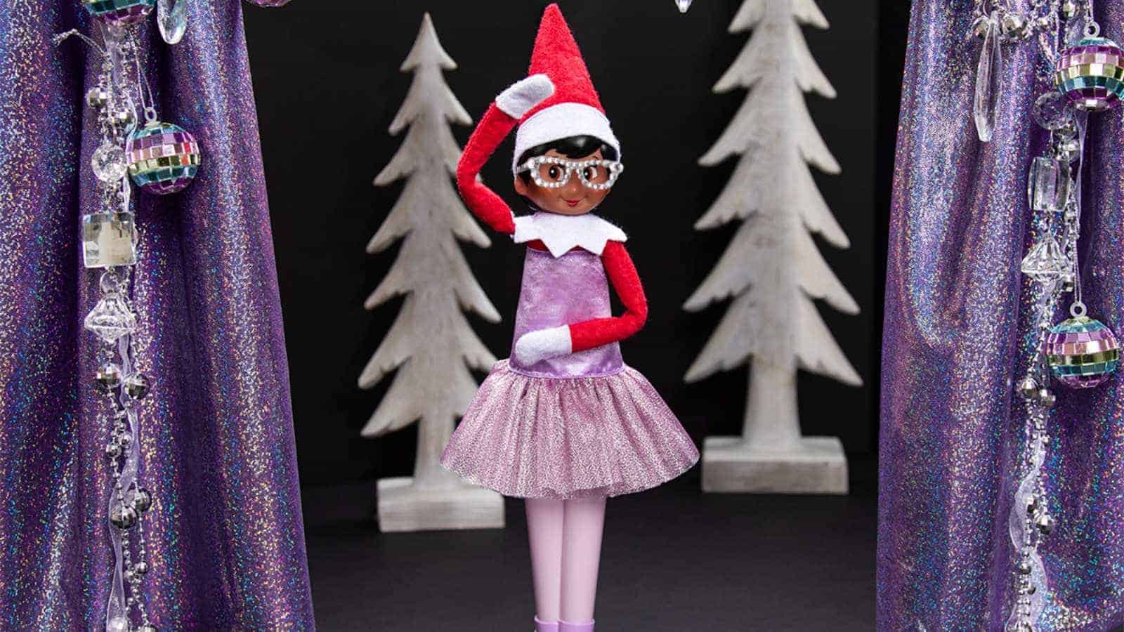 A doll dressed as an elf in a red hat, glasses, and a purple tutu stands in front of white Christmas trees and purple, sparkly decorations.