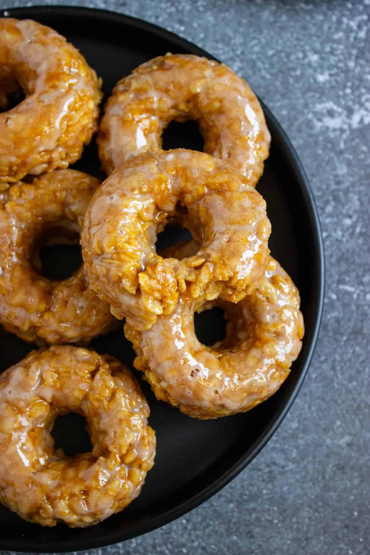 A black plate with several glazed cake donuts stacked and arranged, set on a gray textured surface.
