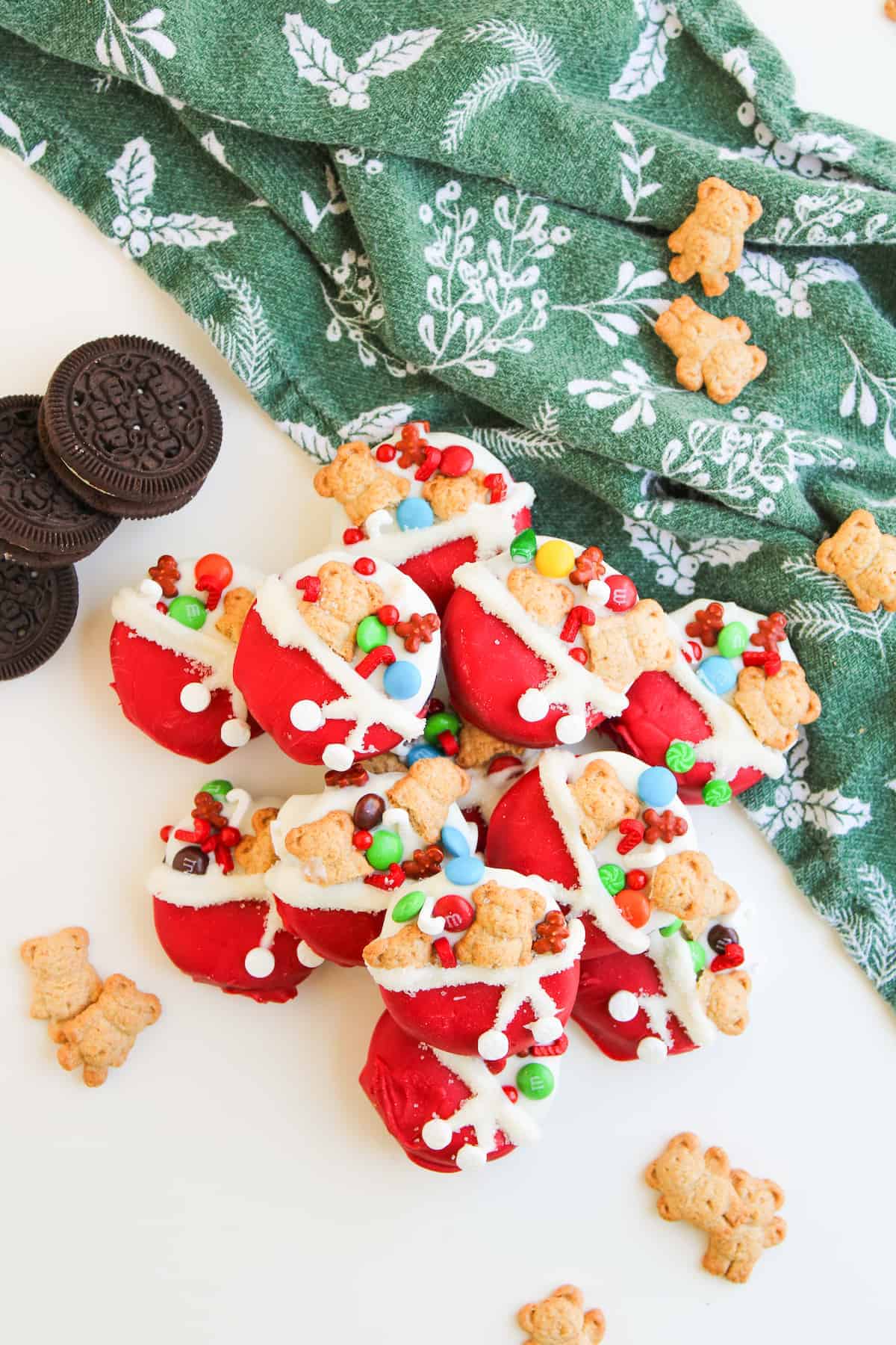 Santa's bag-shaped treats decorated with red icing, teddy bear cookies, and colorful candies are arranged on a white surface with Oreo cookies and a green holiday-patterned cloth.
