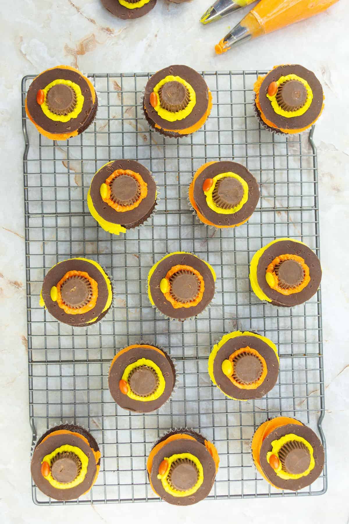 Chocolate cupcakes on a cooling rack, each topped with a peanut butter cup and decorated with yellow and orange icing. Two piping bags with icing are visible in the corner.