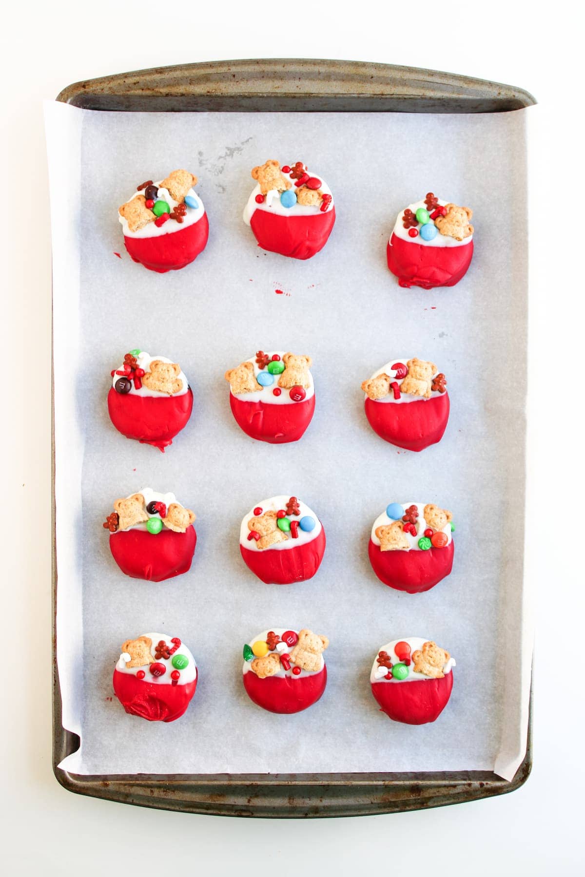 A baking tray lined with parchment paper holds twelve decorated treats dipped in red coating, topped with small candies and Santa's toy bag-shaped cookies.