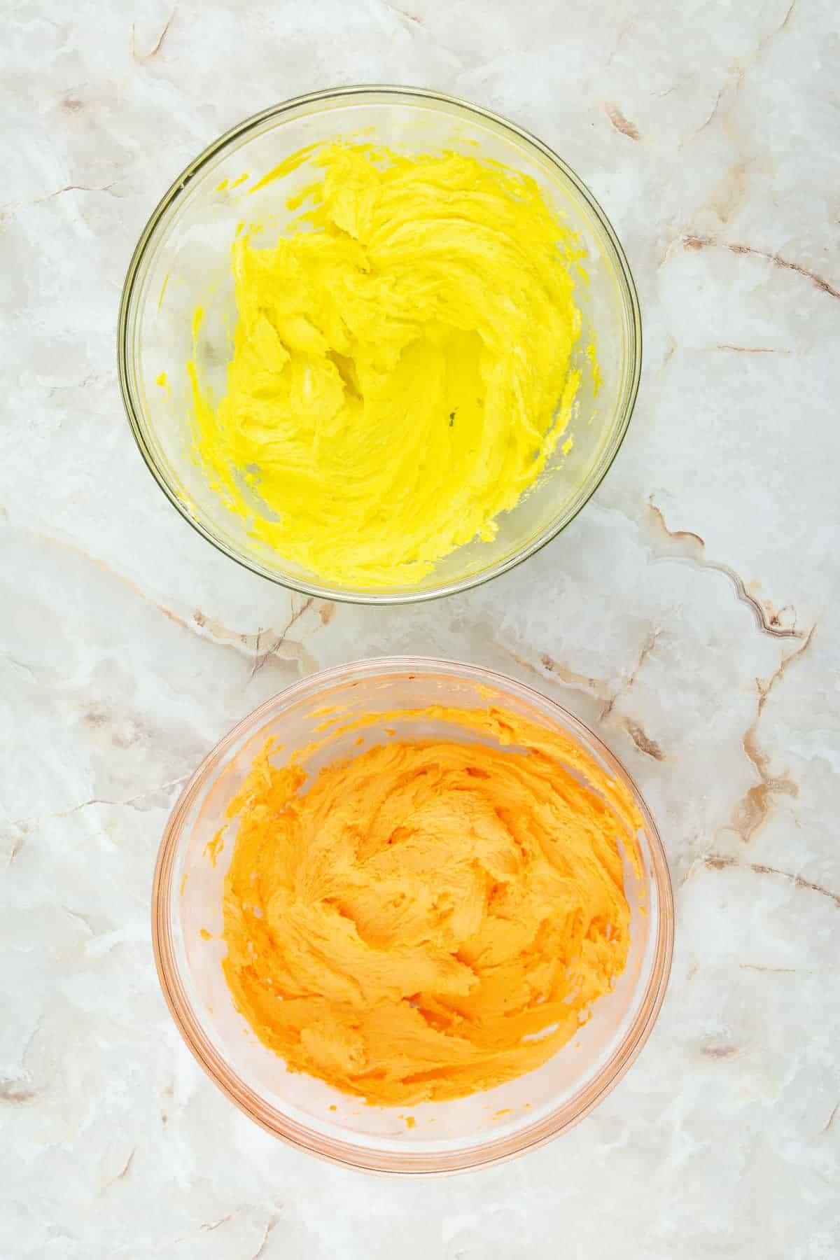 Two glass bowls on a marble surface; the top bowl contains yellow creamy mixture, and the bottom bowl contains an orange creamy mixture.