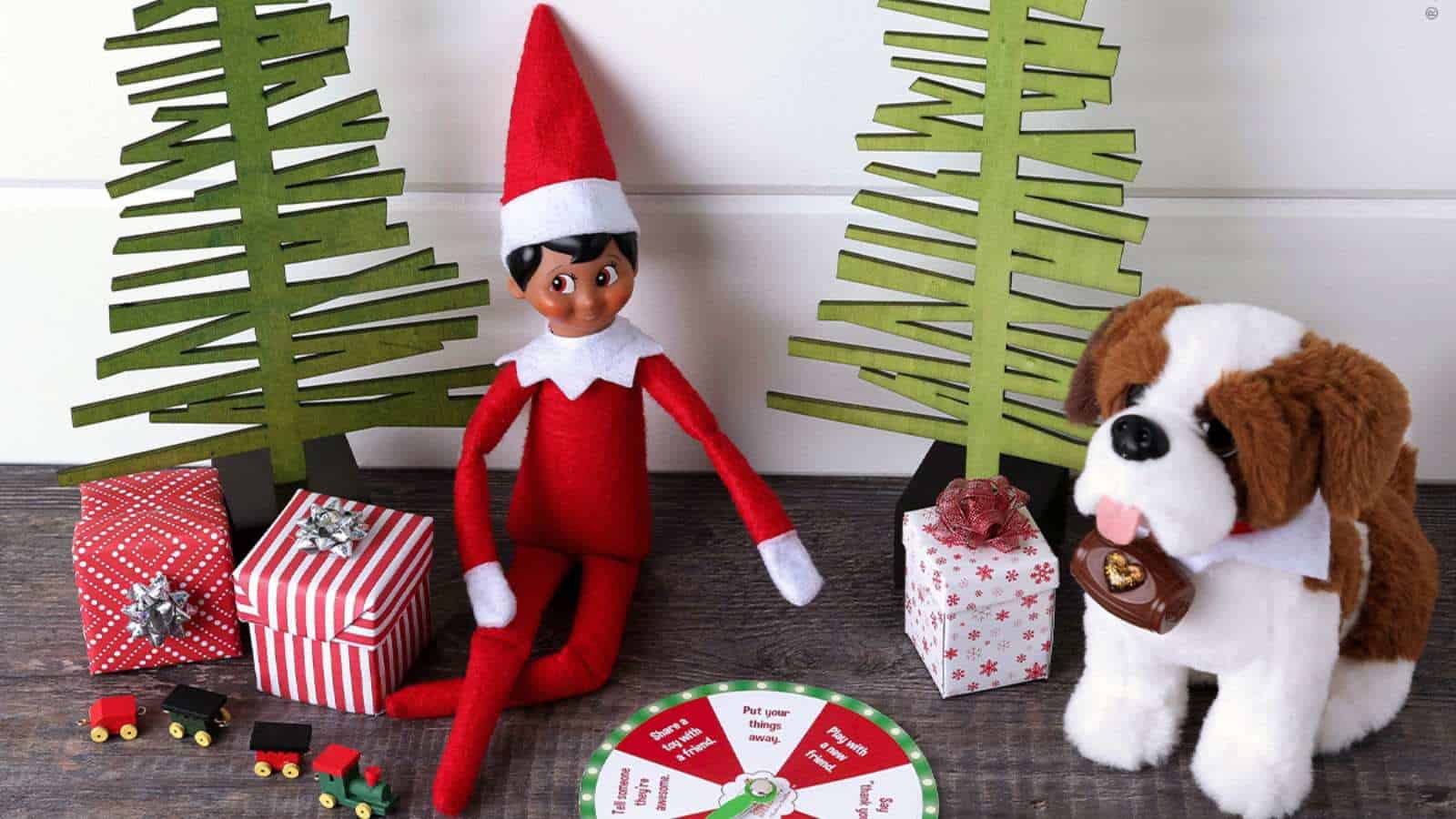 A plush elf doll sits between two toy trees, surrounded by wrapped gifts, a toy dog, a small train, and a circular holiday-themed spinner on a wooden surface.