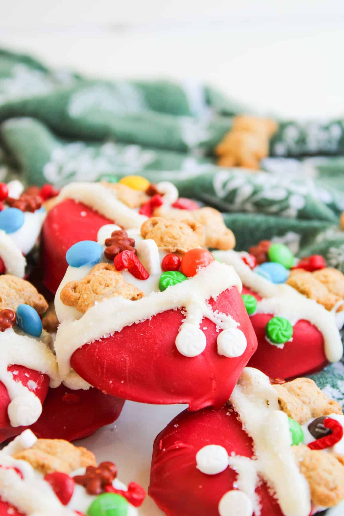 Cookies decorated to look like Santa’s toy bag, with red icing, white frosting trim, sprinkles, and small candies, are arranged on a surface near a green holiday-patterned cloth.