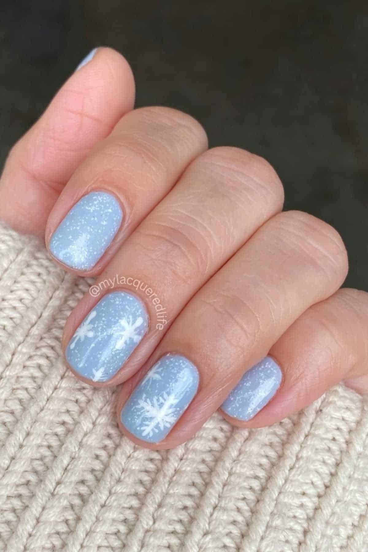 A hand with short, light blue nails featuring white snowflake designs and small white speckles, resting on a cream knit sweater.