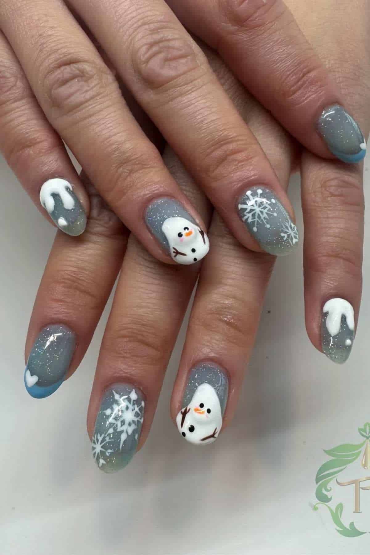 Hands with grey-blue, glittery winter-themed nail art featuring white snowflakes, a snowman design, dripping snow tips, and a small heart accent.