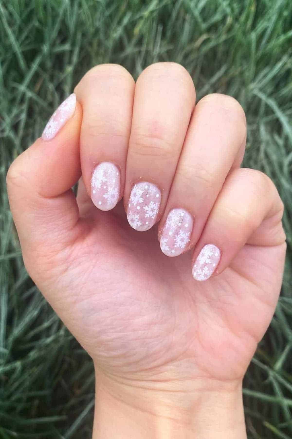 A hand with short nails featuring pale pink nail polish and white snowflake designs, shown against a blurred green grass background.