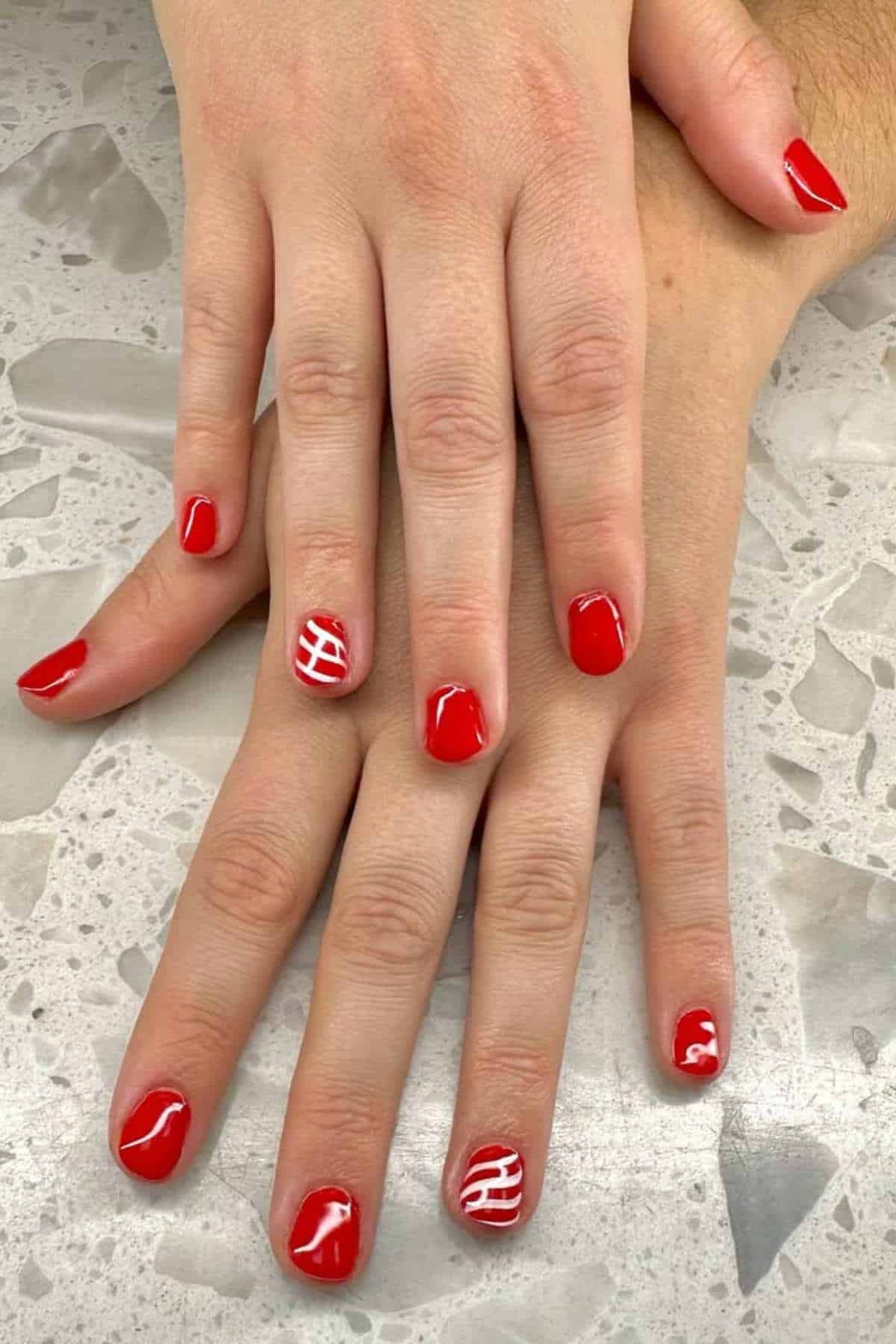 Two hands with short, red-painted nails; each ring finger features a white, striped line pattern. The hands rest on a light, textured surface.