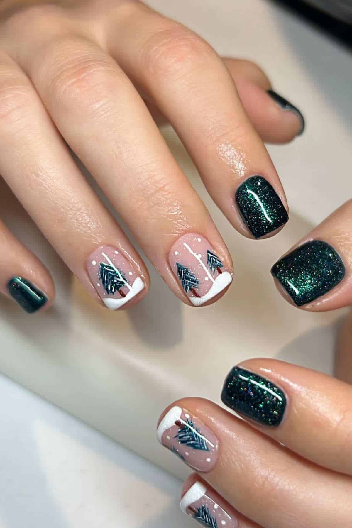 Close-up of hands with dark green glitter nail polish and accent nails featuring white tips and small painted green tree designs.