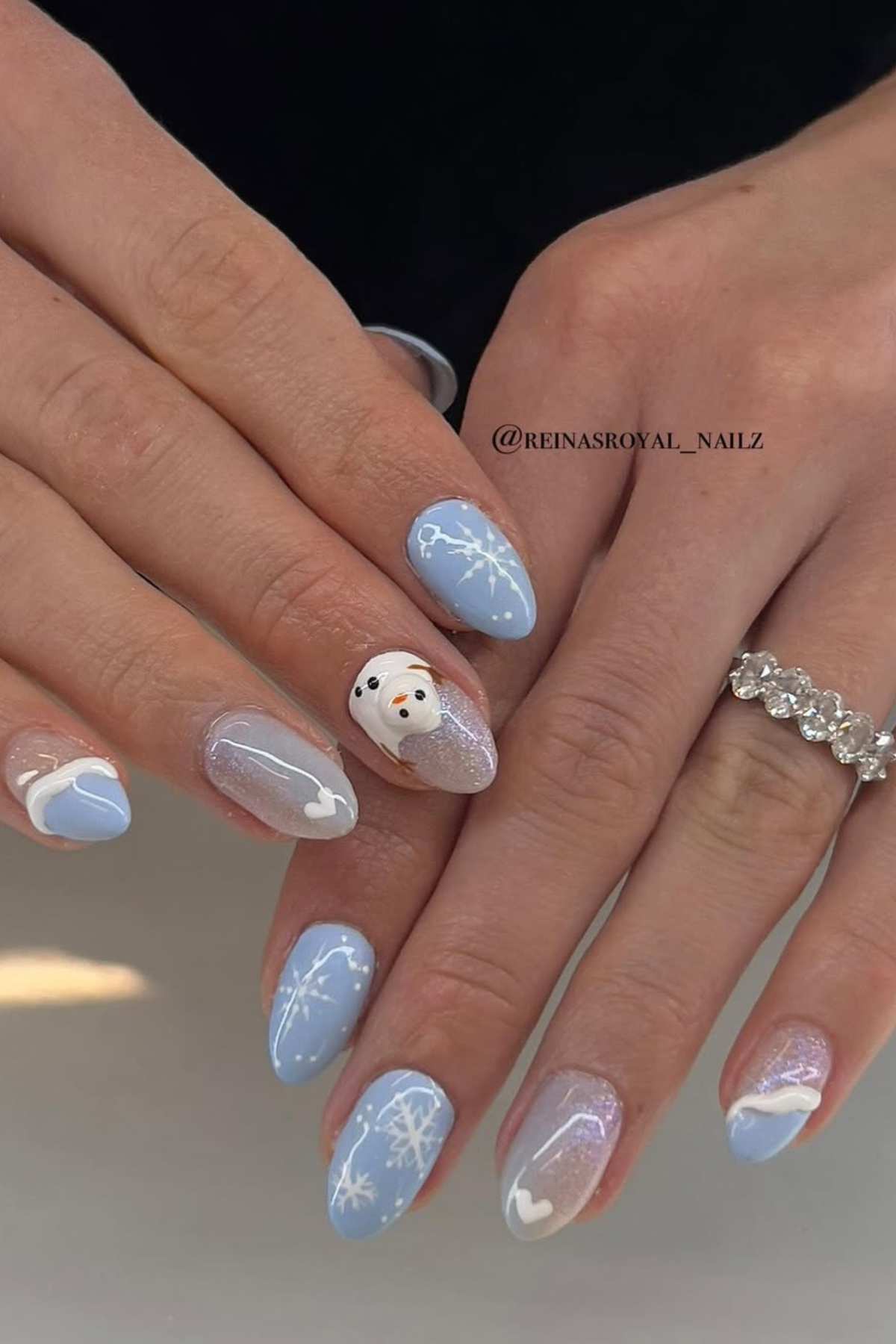 Hands with almond-shaped nails painted in light blue and white winter-themed designs, featuring snowflakes, hearts, and a snowman accent nail.