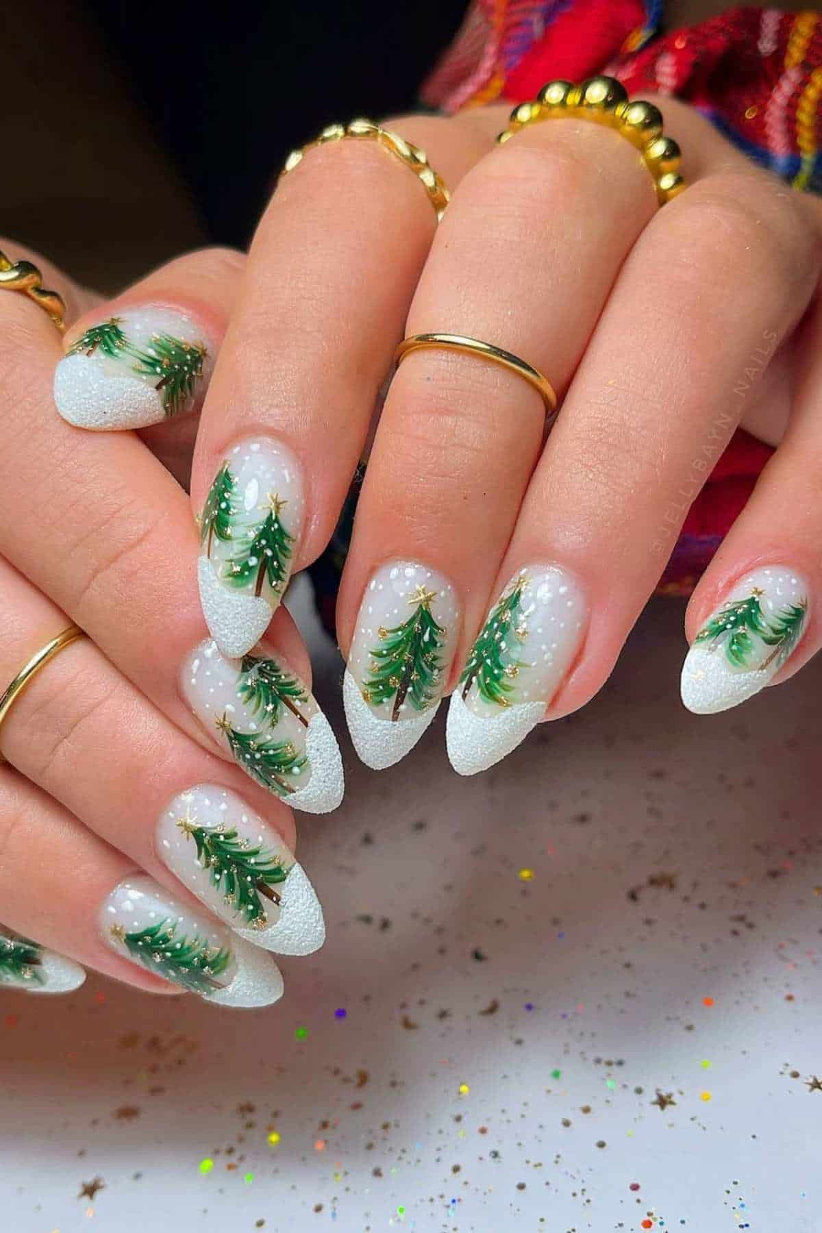Hands with long, almond-shaped nails featuring Christmas tree designs and white, glittery tips. The person is wearing gold rings, and there are colorful sparkles on the surface below.