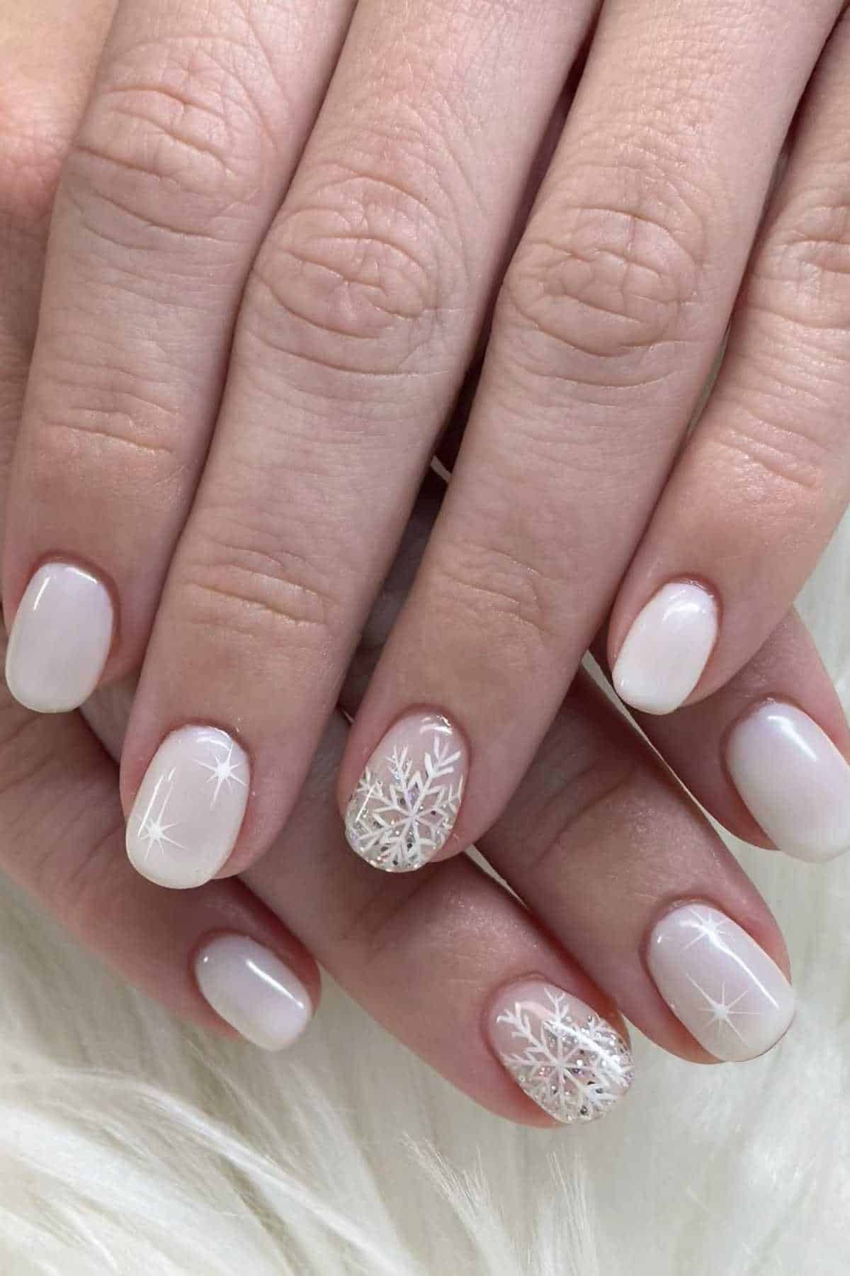 Hands with short, oval-shaped nails painted white and decorated with snowflake designs and subtle shimmer accents.