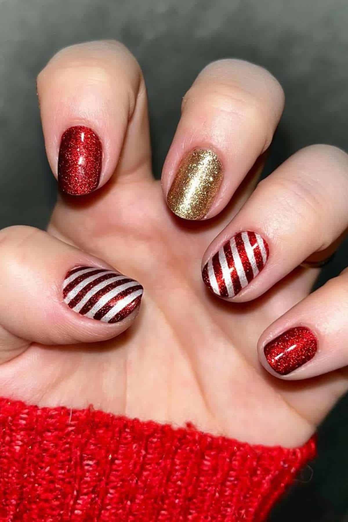 A hand with festive nail art featuring red glitter, gold glitter, and red-and-white candy cane stripes. The person is wearing a red knit sleeve.
