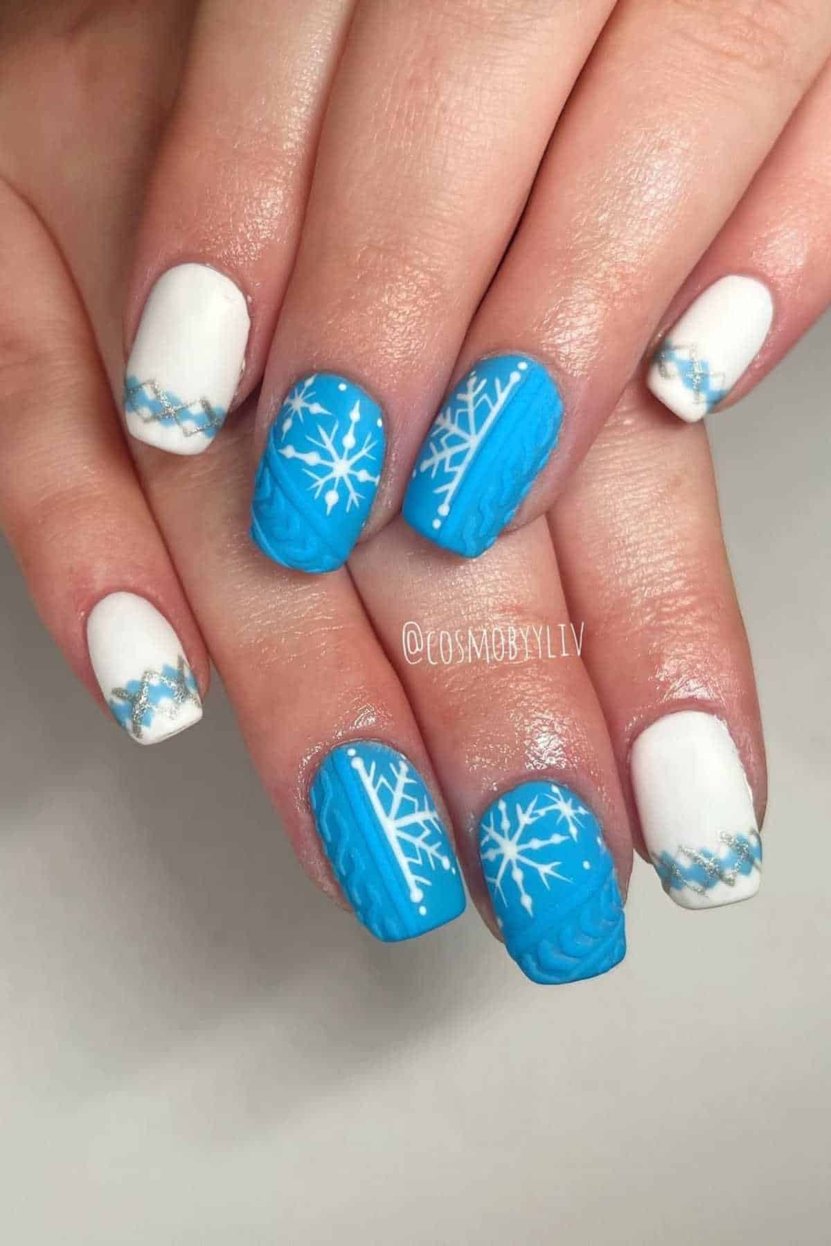 Close-up of hands with winter-themed nail art; nails are painted blue and white with snowflake designs and knit pattern accents.