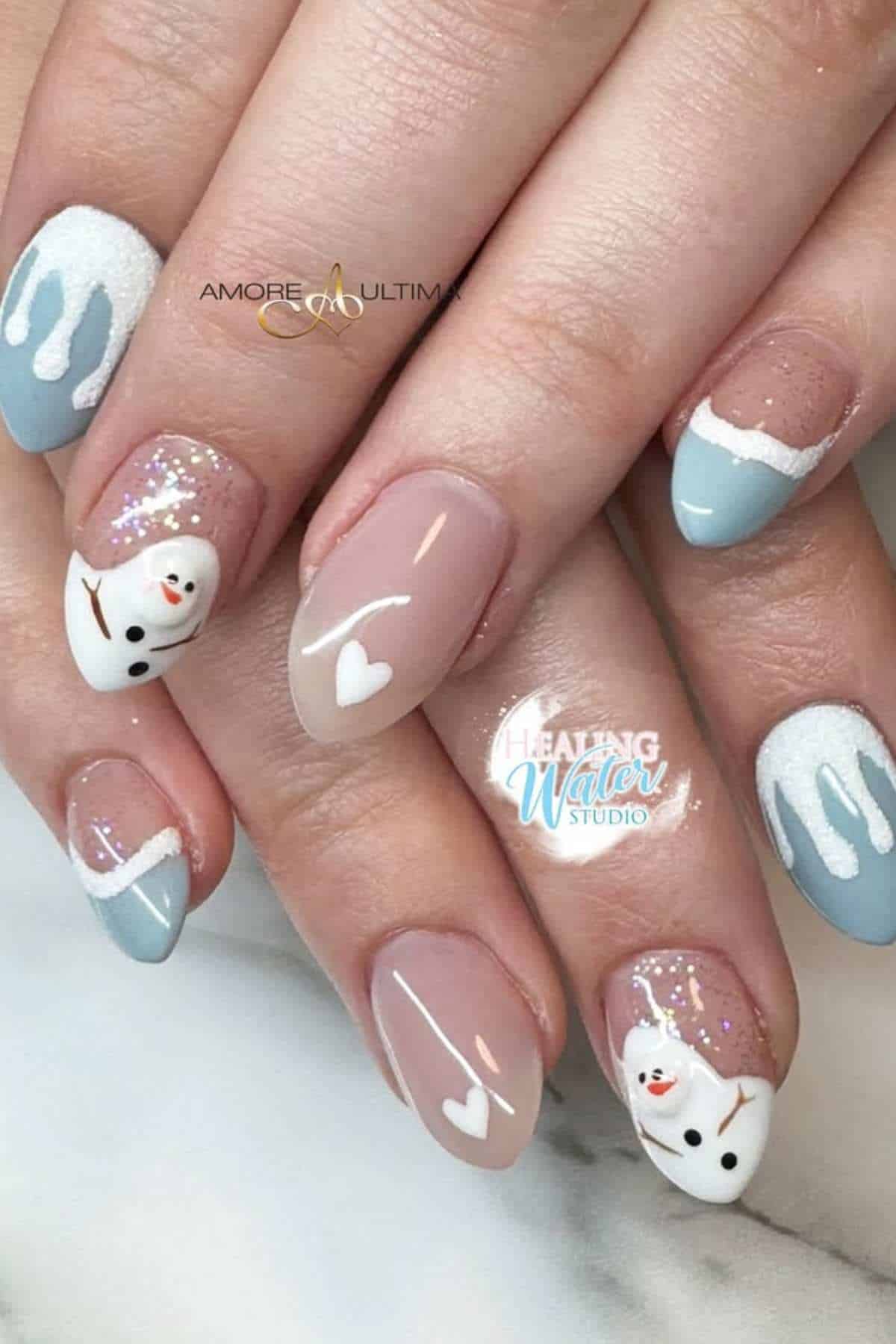 Hands with almond-shaped nails featuring winter-themed nail art: blue and white tips, glitter, white hearts, and snowman designs on two nails.
